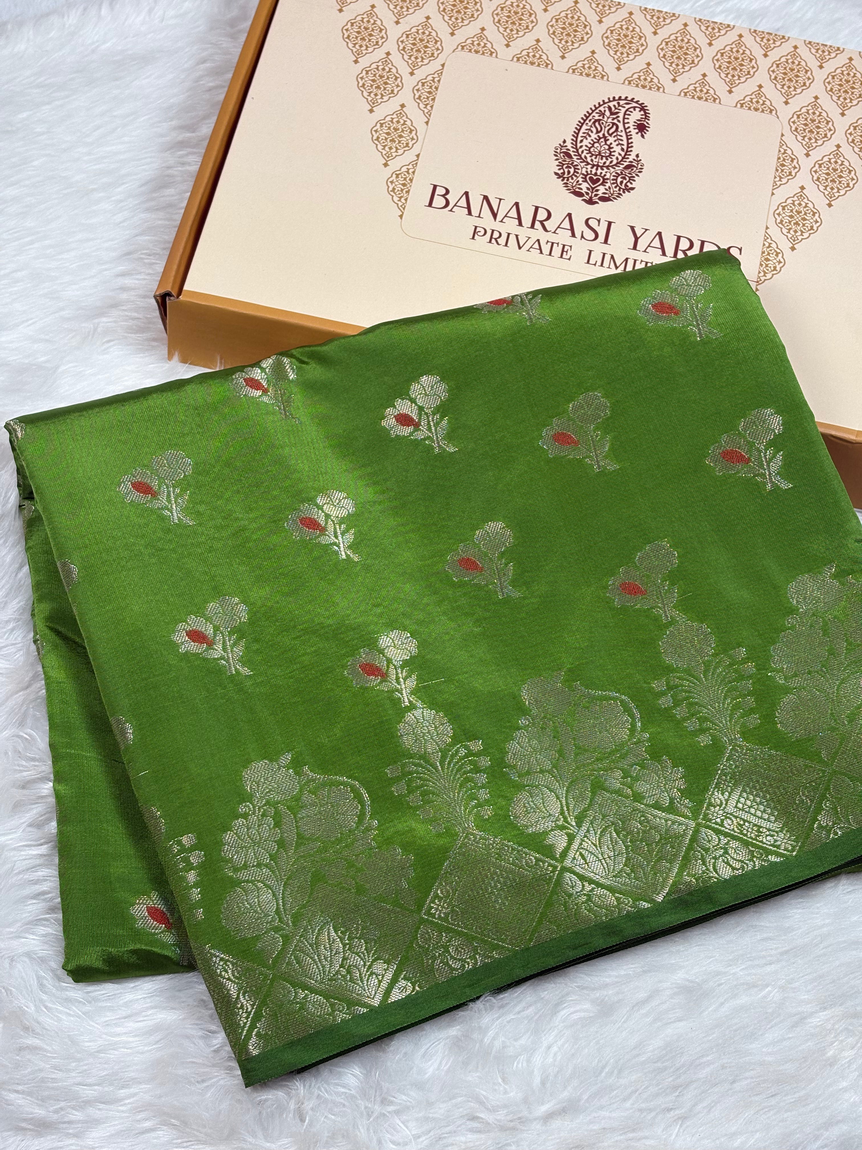 A folded green silk saree with silver floral motifs and small mina accents, displayed next to its Banarasi Yards box on a white faux fur surface.