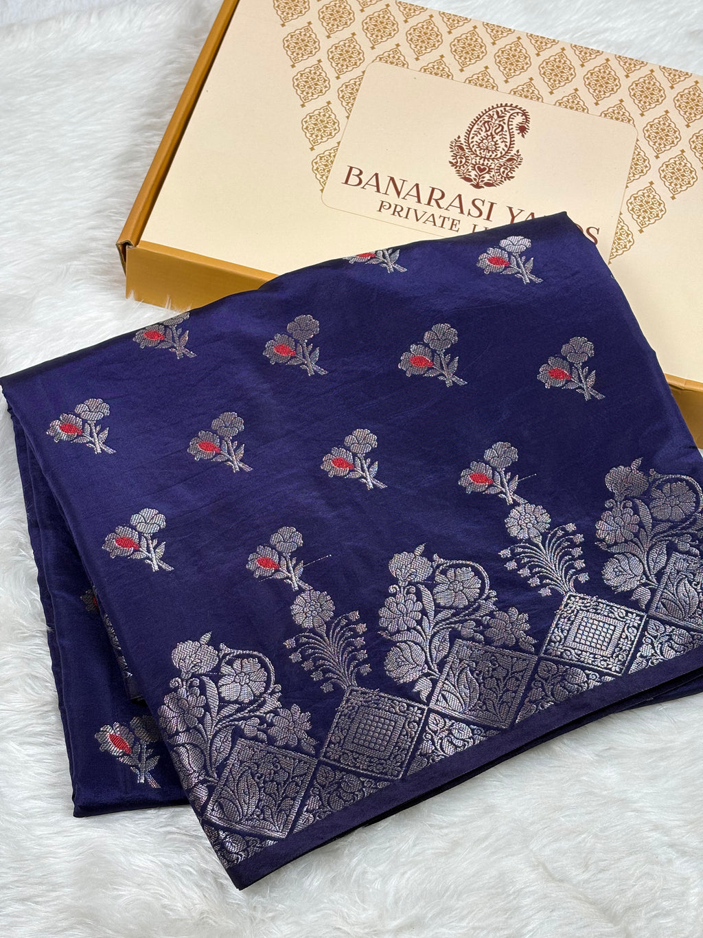 A folded navy blue Banarasi silk saree with silver floral motifs and small mina accents, displayed next to its Banarasi Yards gift box on a white faux fur background.
