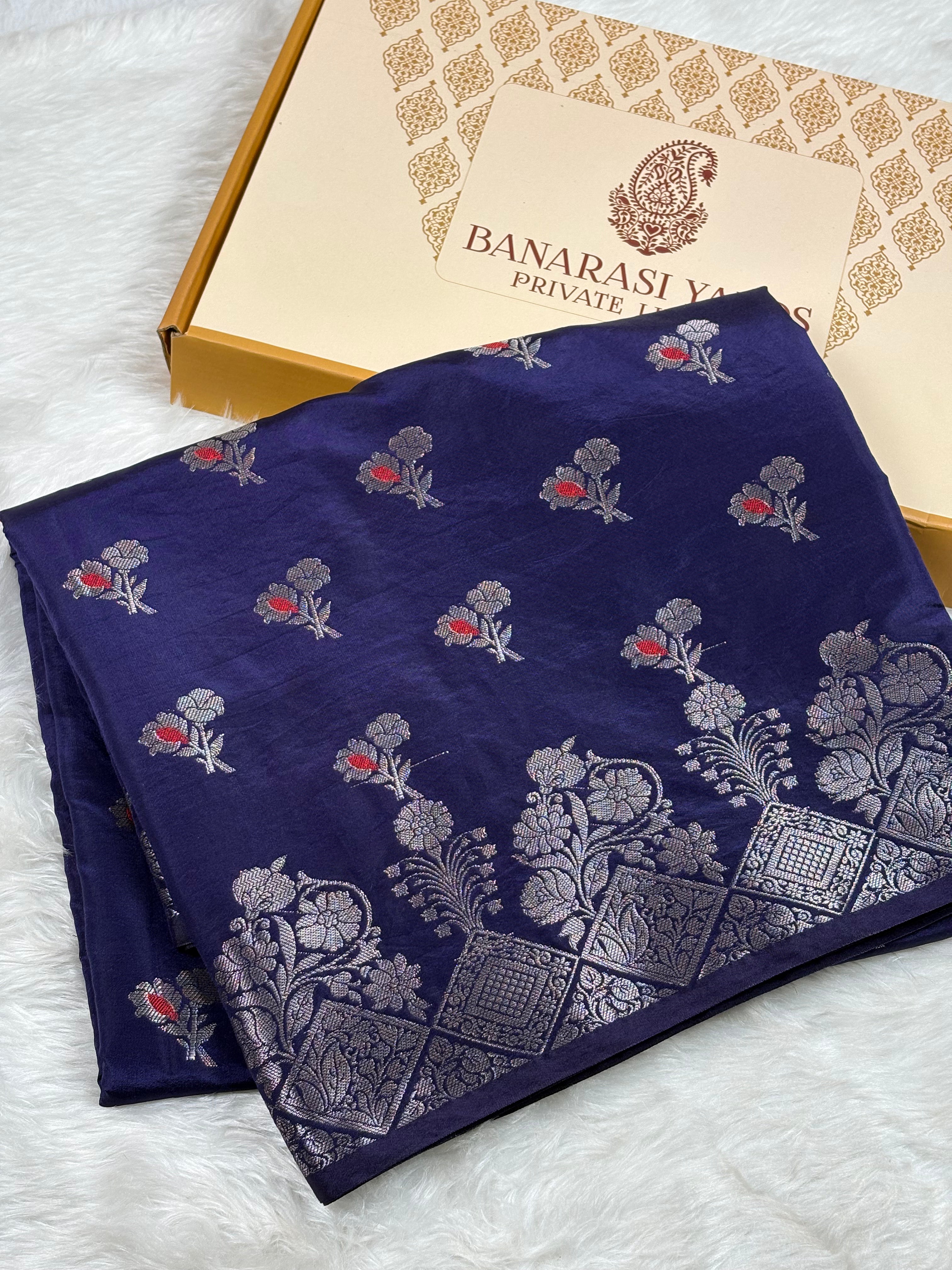 A folded navy blue Banarasi silk saree with silver floral motifs and small mina accents, displayed next to its Banarasi Yards gift box on a white faux fur background.