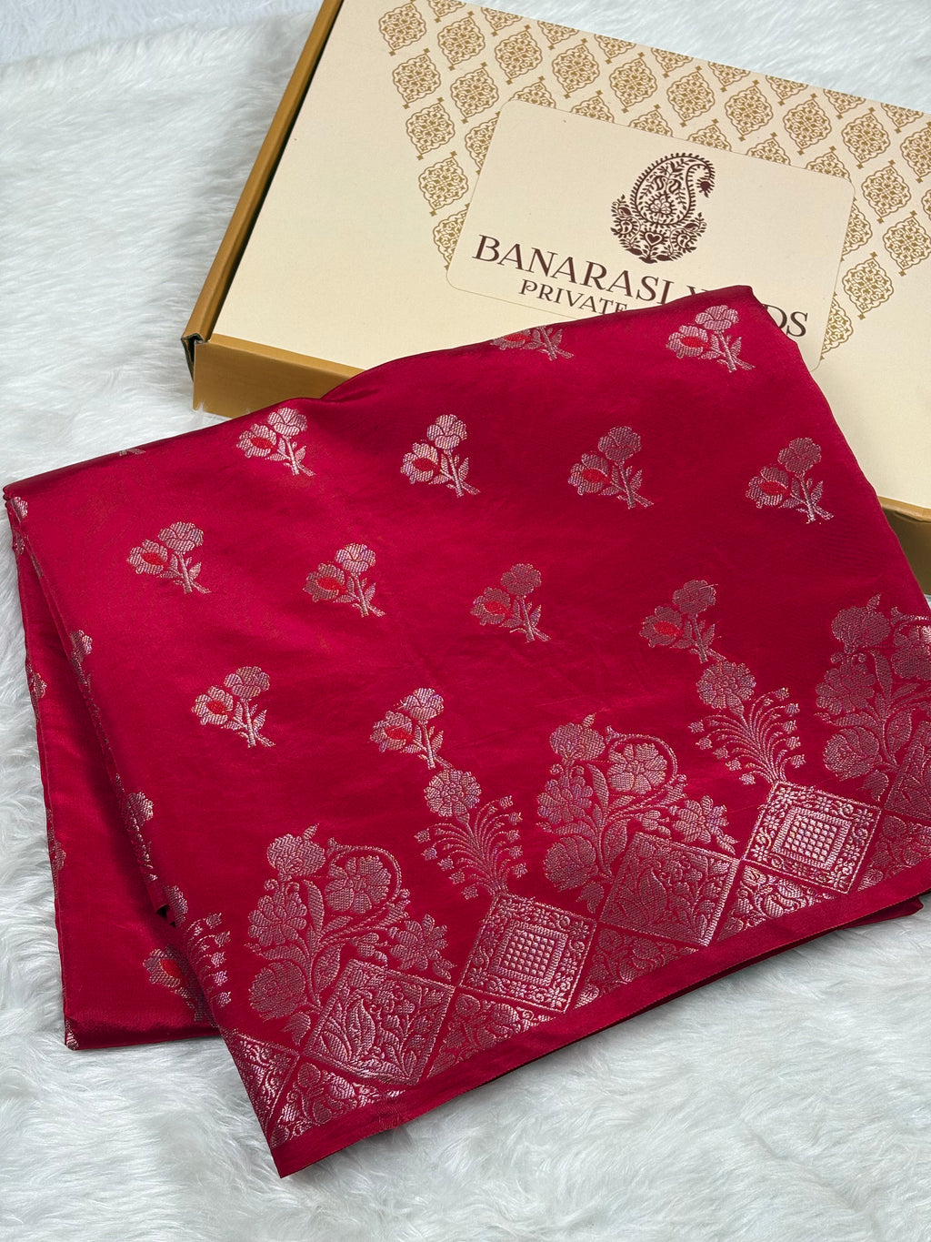 A folded deep red silk saree with gold floral and geometric patterns, displayed next to its Banaras Yards Private Limited box on a white faux fur background.