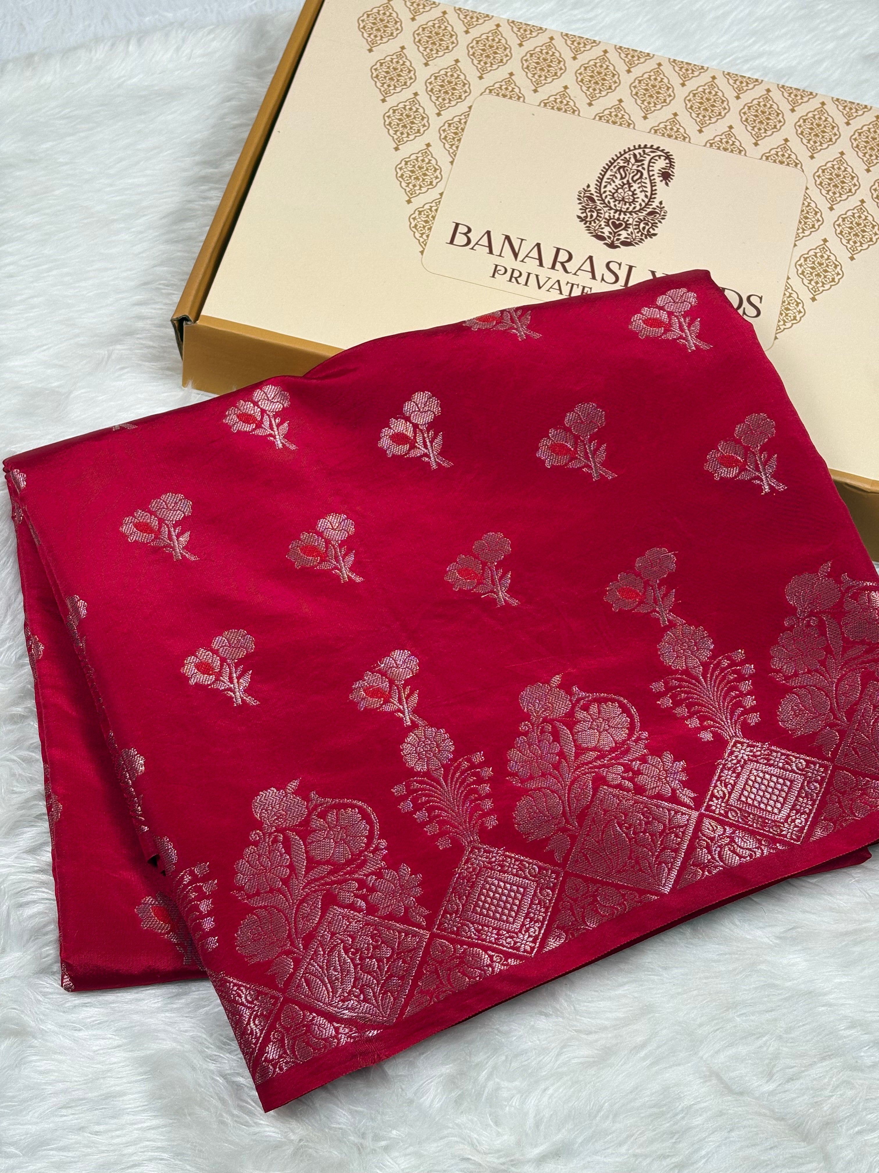 A folded deep red silk saree with gold floral and geometric patterns, displayed next to its Banaras Yards Private Limited box on a white faux fur background.