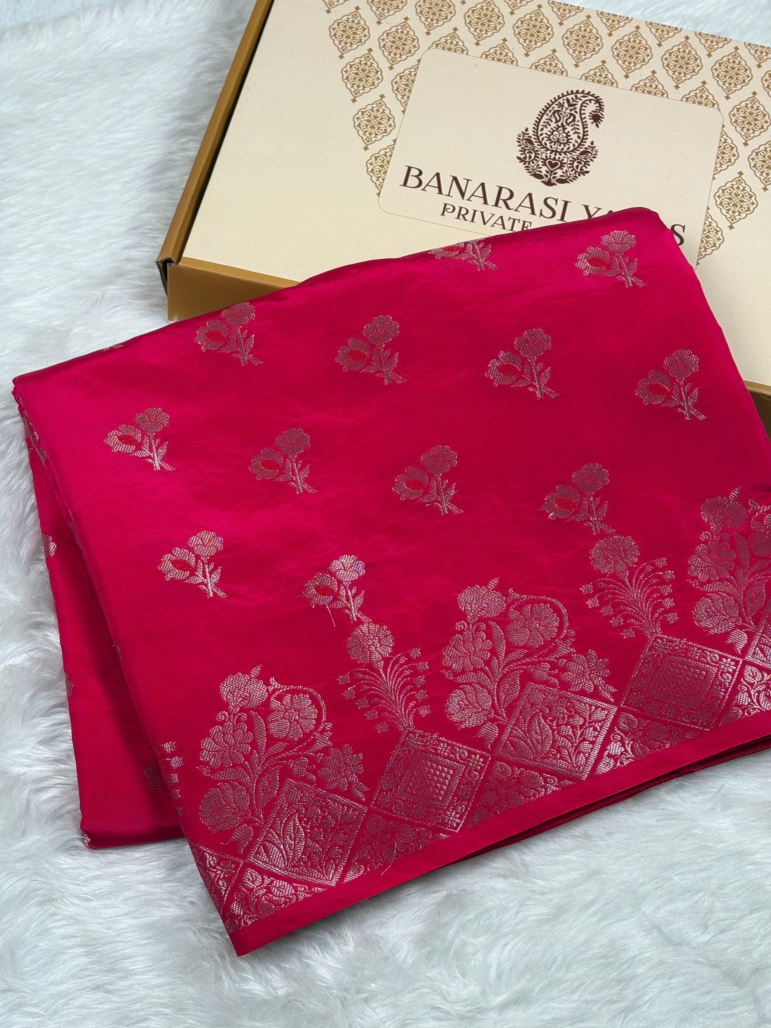 A folded bright pink saree with gold floral mina buti and geometric motifs, displayed next to a branded Banarasi Yards Private Limited box on a white faux-fur surface.