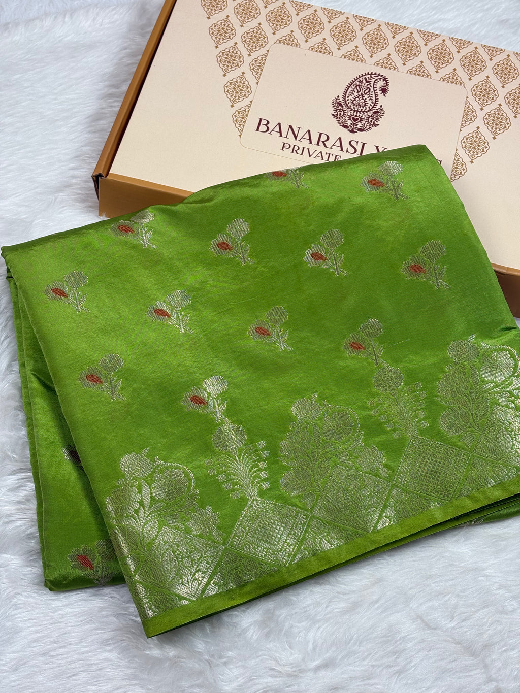A folded green Banarasi silk saree with gold floral motifs and mina accents, displayed next to its Banarasi Yards Private Limited brand box on a white textured background.