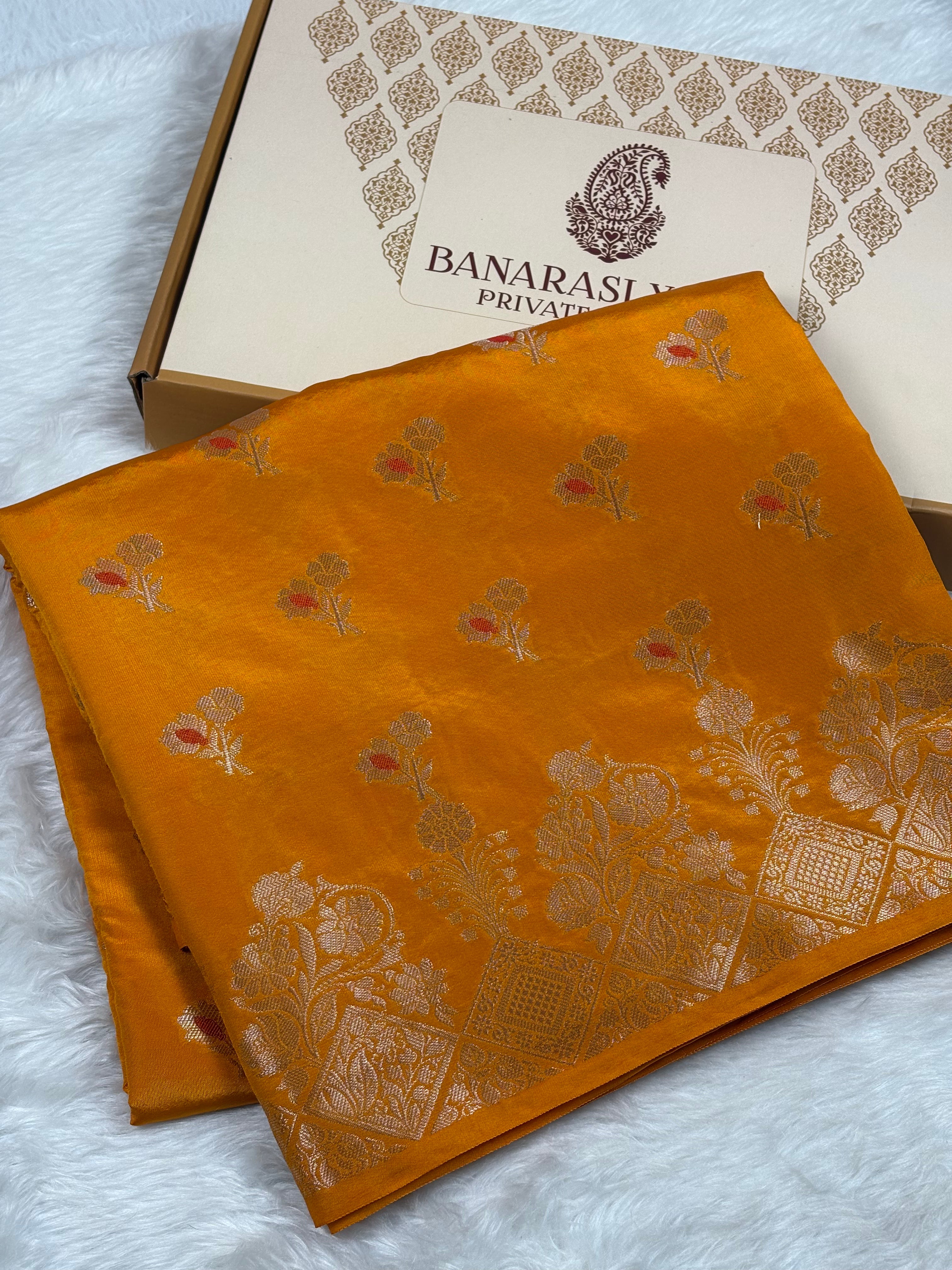 A folded mustard yellow Banarasi silk saree with intricate gold floral mina motifs, displayed next to its branded packaging box on a white faux fur surface.