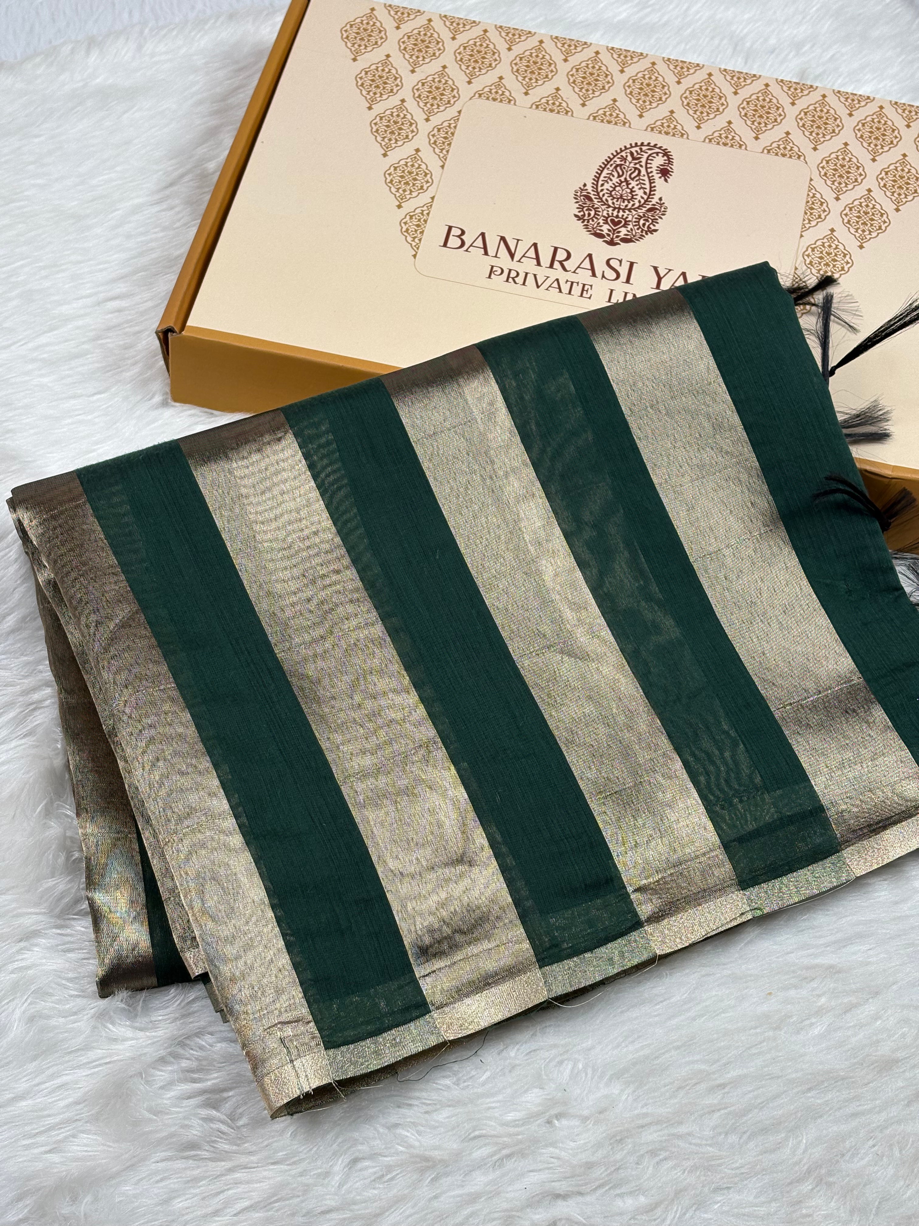 A folded Banarasi saree with dark green and gold stripes, displayed next to its branded packaging box on a white faux fur surface.