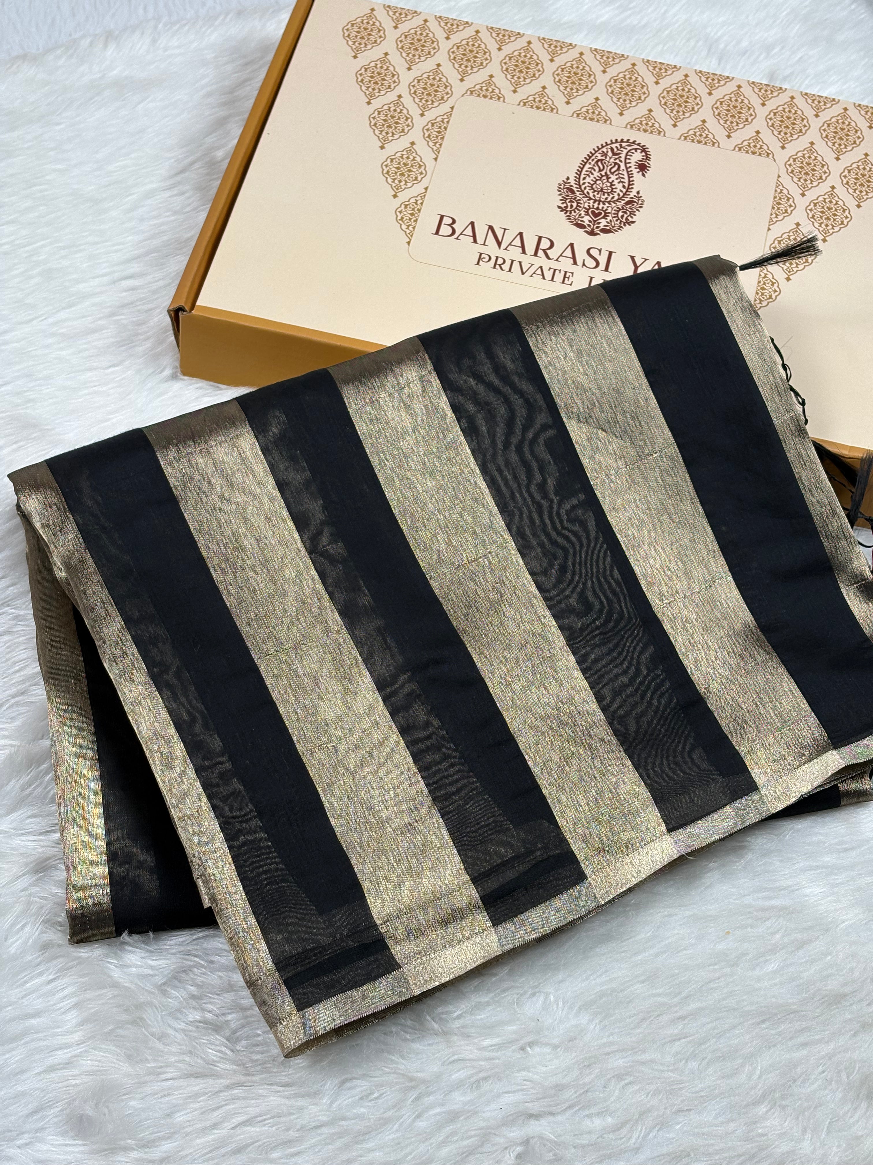 A folded Banarasi saree with black and gold stripes, displayed next to its branded packaging box on a white faux fur surface.