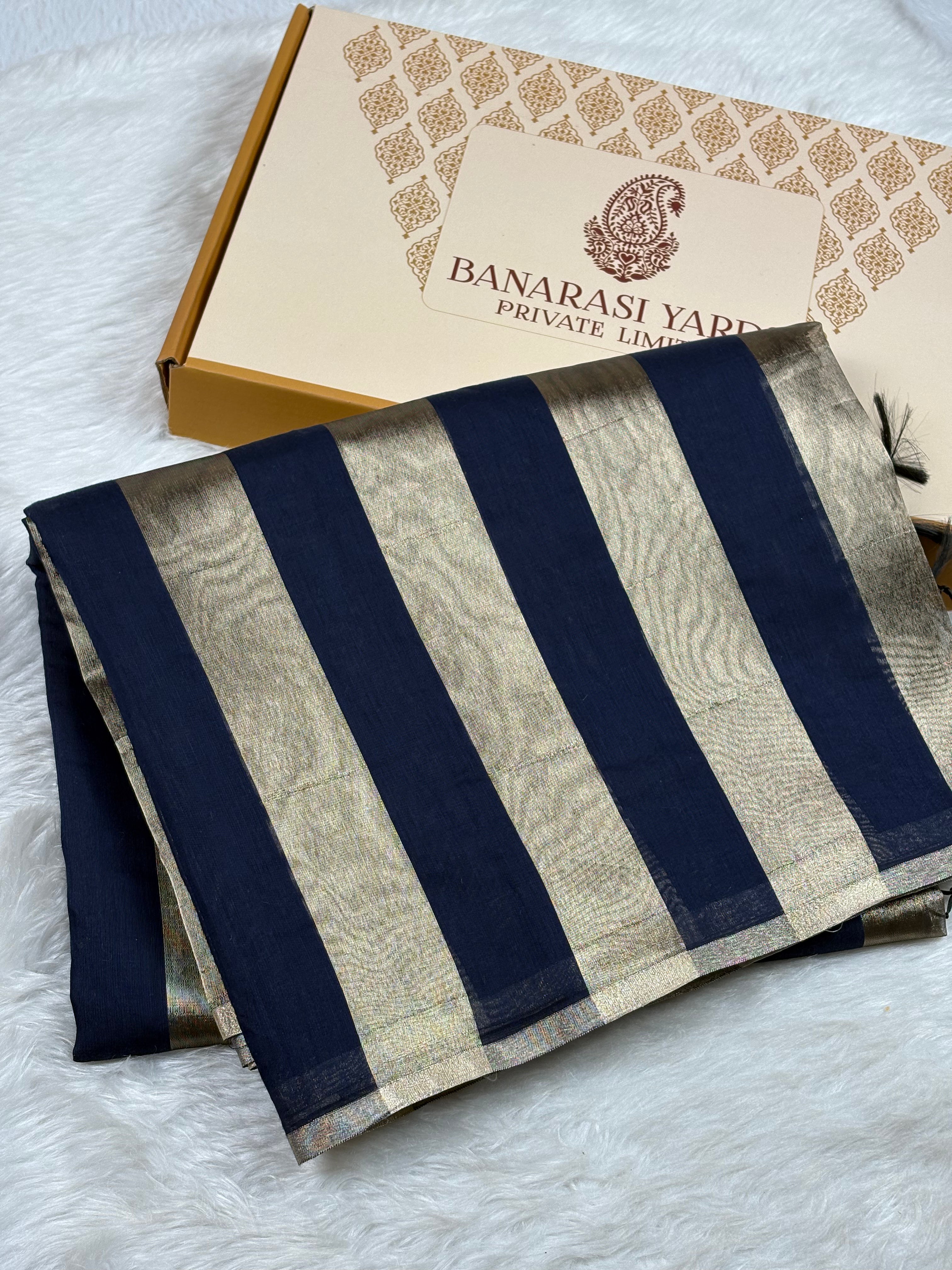 A folded Banarasi saree with navy blue and gold stripes, displayed next to its branded packaging box on a white faux fur surface.