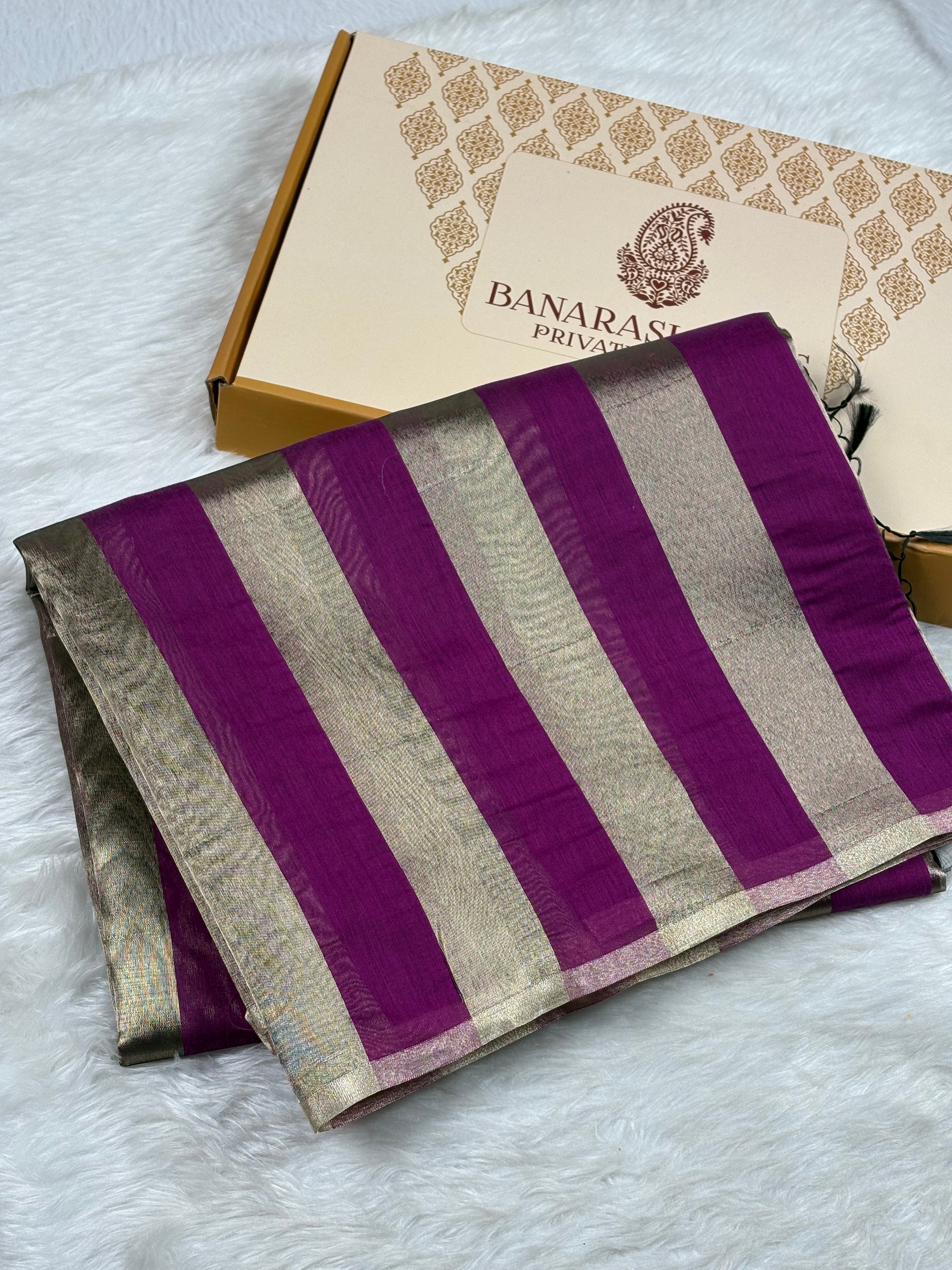 A folded purple and gold striped saree with zari borders, displayed next to its branded Banarasi Yards Private Limited box on a white faux fur surface.