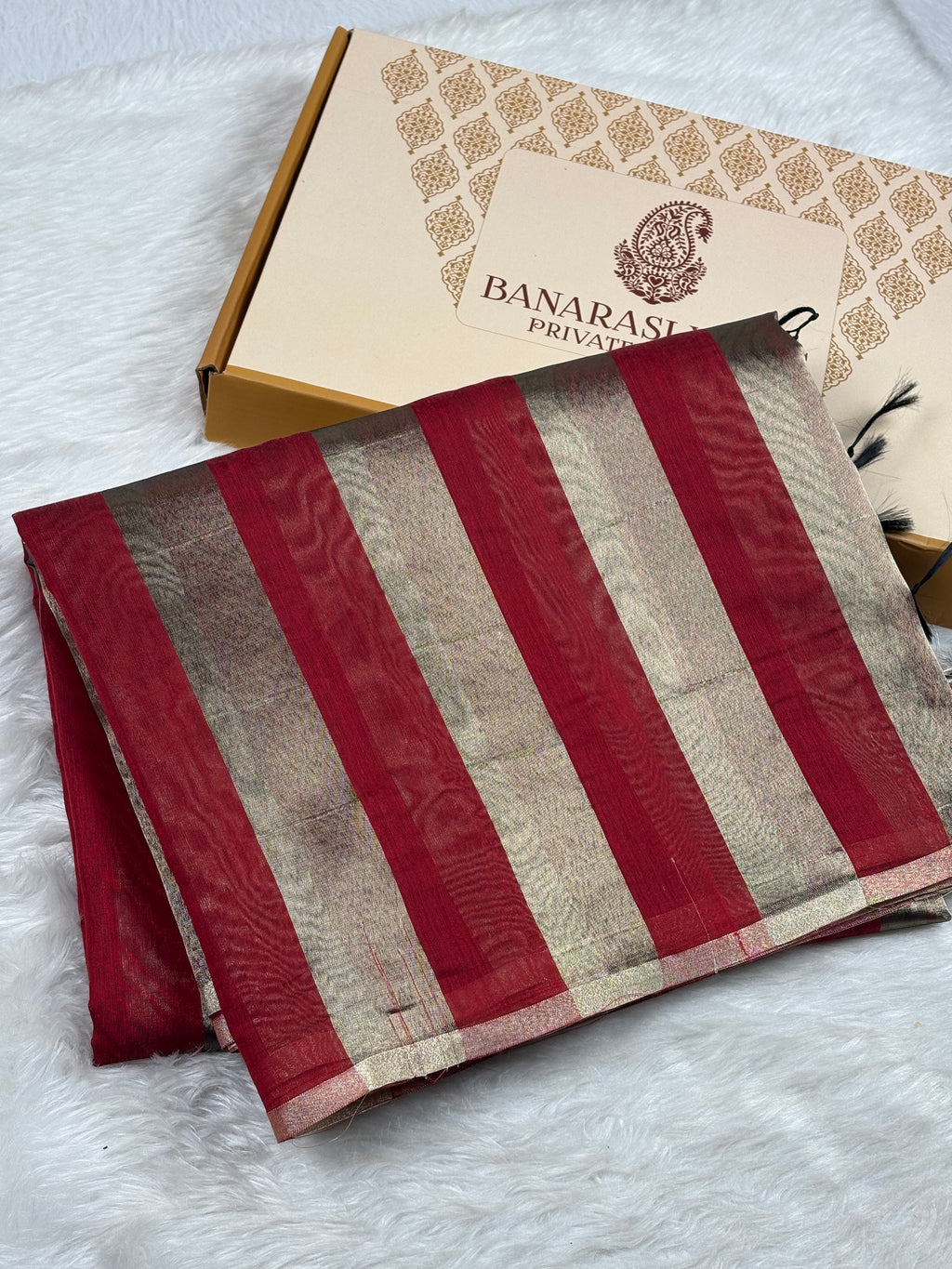 A folded Banarasi saree with vertical stripes of deep red and gold, displayed next to its branded packaging box on a white faux fur surface.