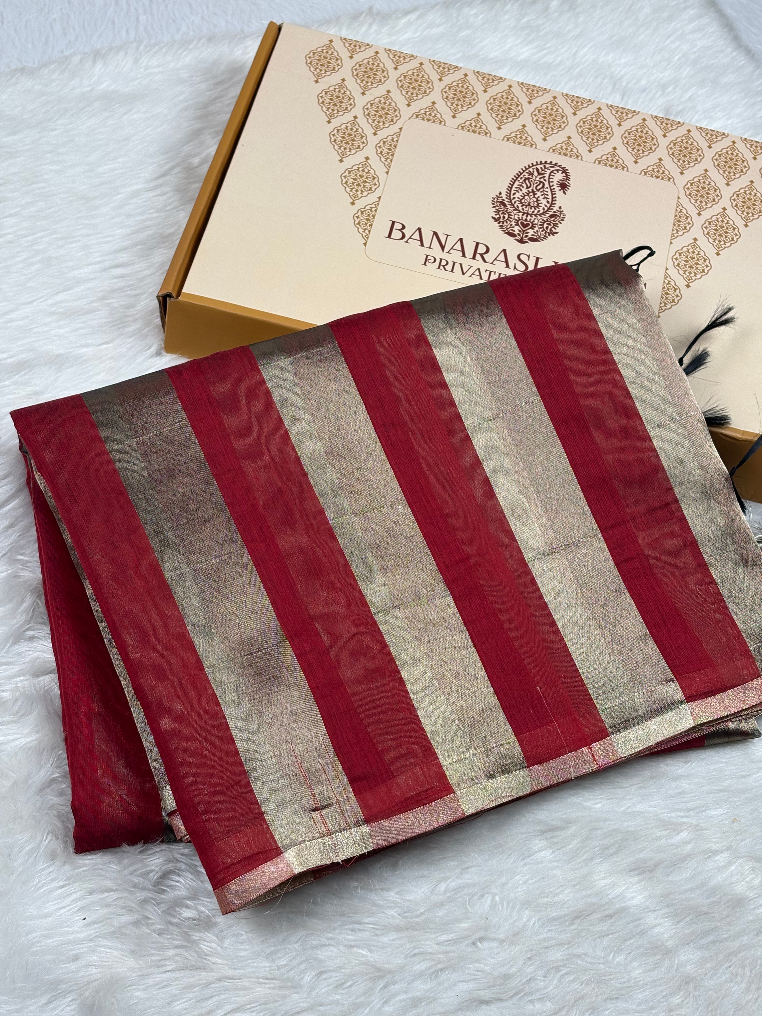 A folded Banarasi saree with vertical stripes of deep red and gold, displayed next to its branded packaging box on a white faux fur surface.