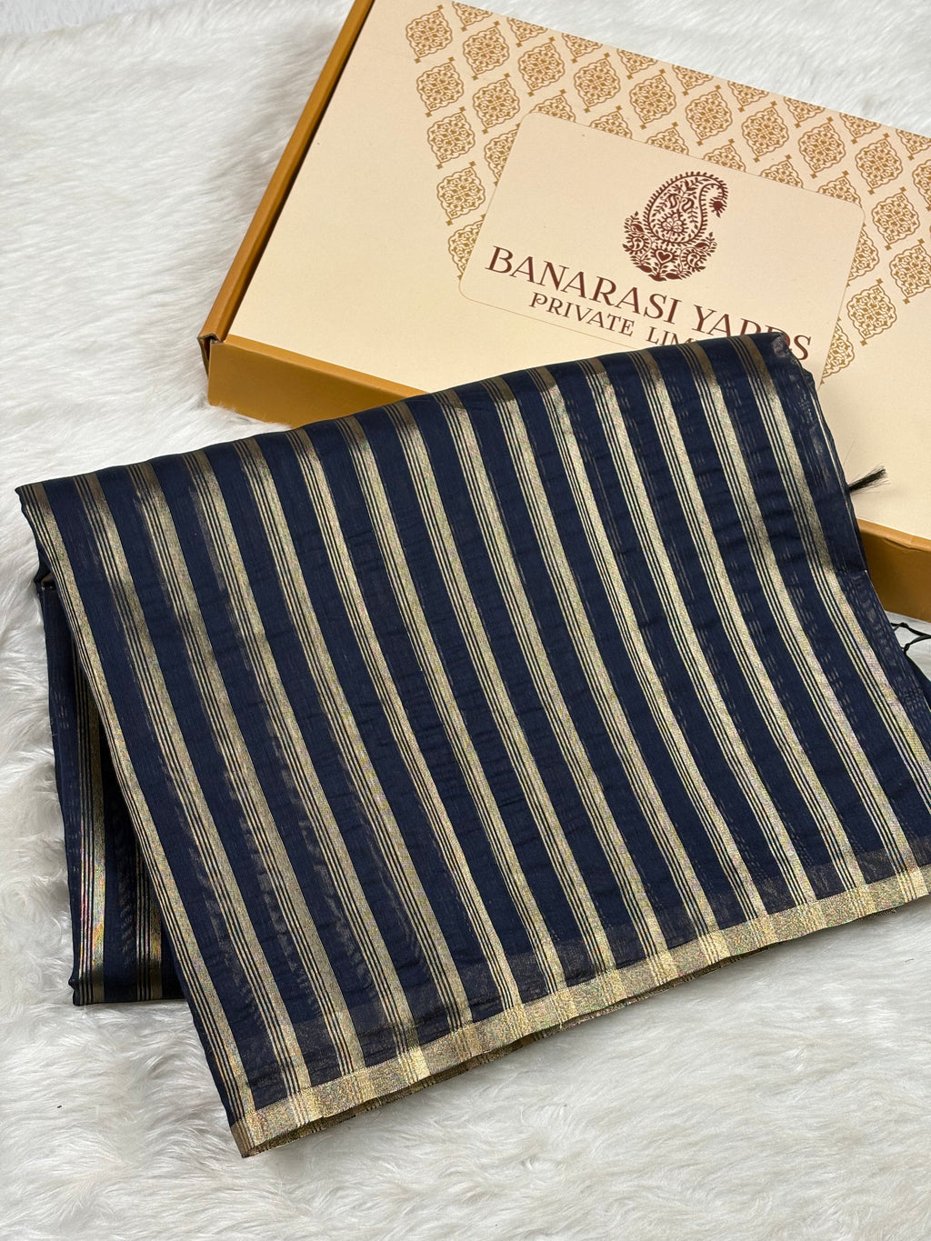 A folded navy blue and gold striped saree, displayed next to its branded packaging box on a white faux fur surface. The box is features the text 'BANARASI YARDS PRIVATE LIMITED'.