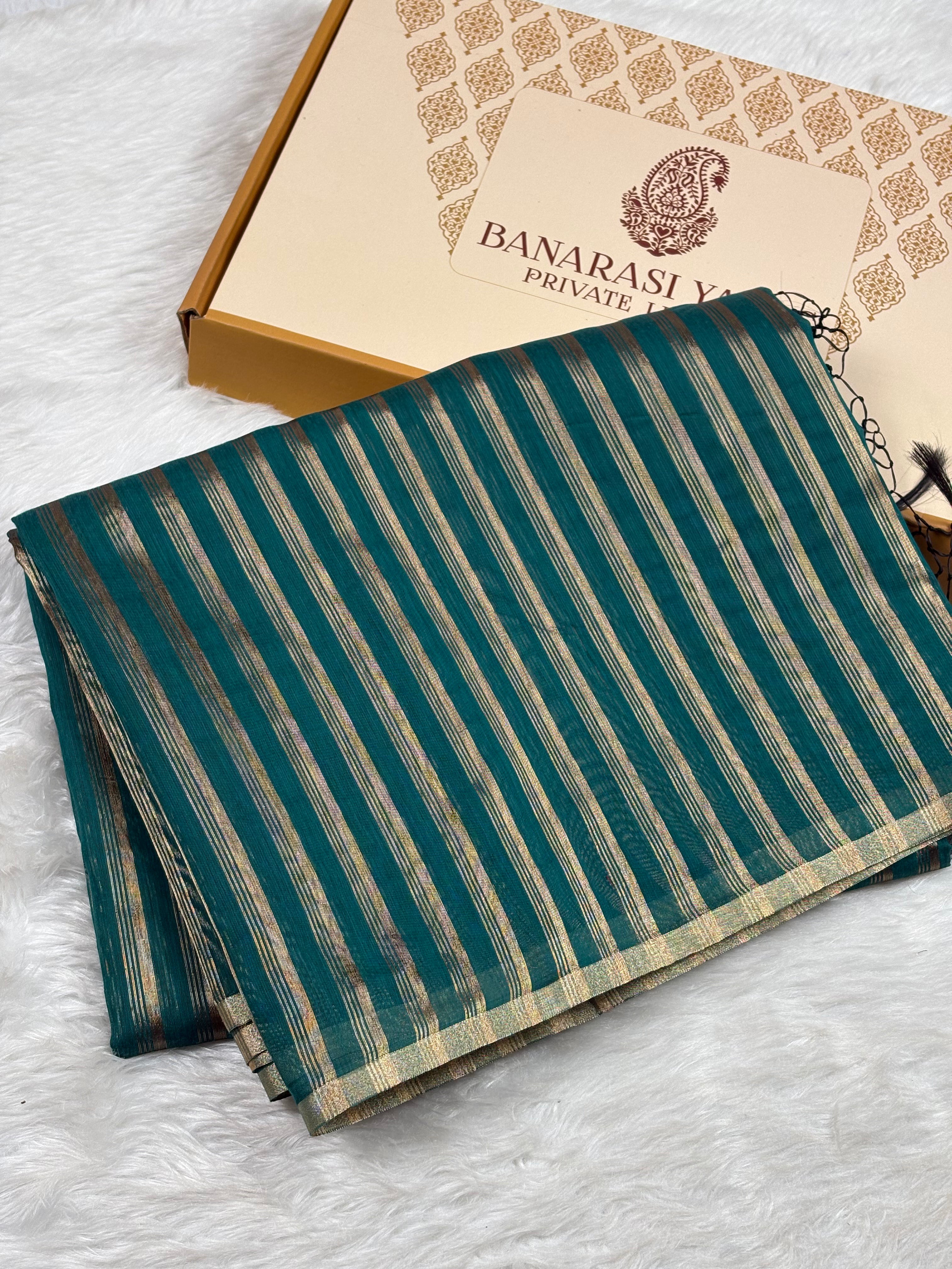 A folded teal green and gold striped saree with gold zari detailing, displayed next to its branded gift box on a white faux fur surface.