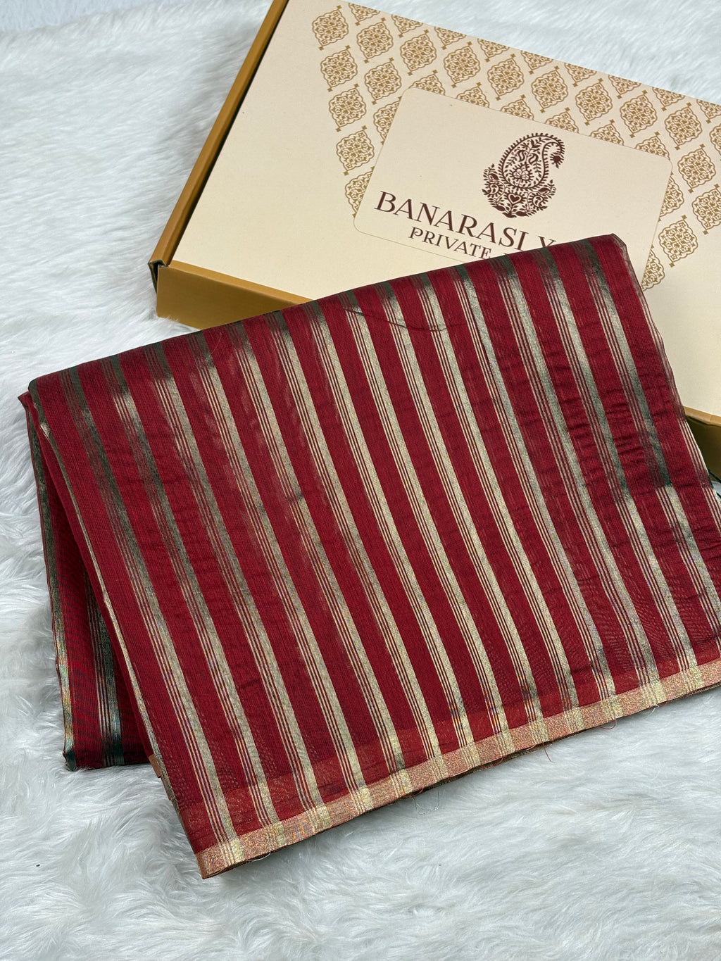 A folded red and gold striped Banarasi saree with zari details, displayed next to its branded packaging box on a white faux fur surface.