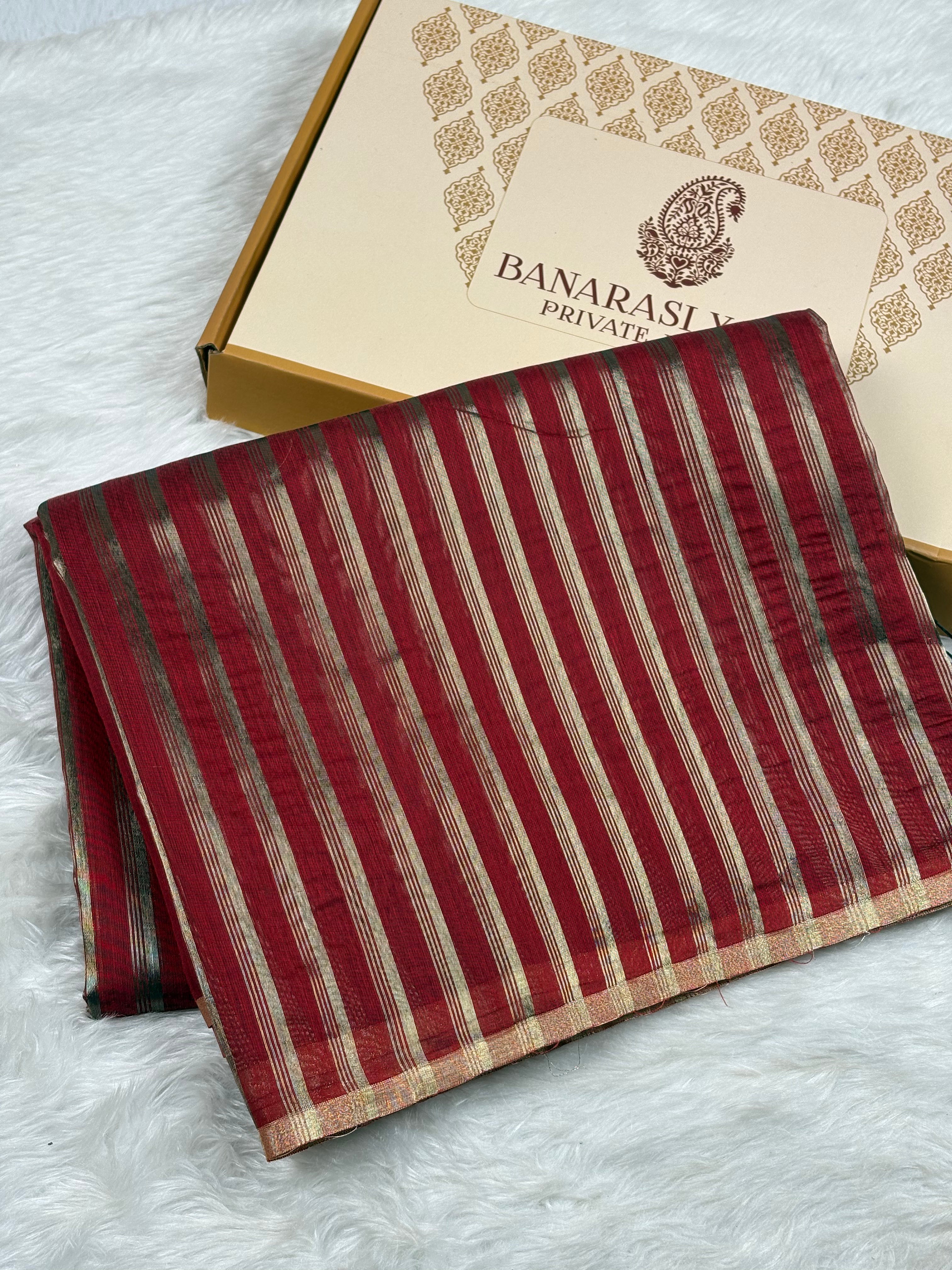 A folded red and gold striped Banarasi saree with zari details, displayed next to its branded packaging box on a white faux fur surface.