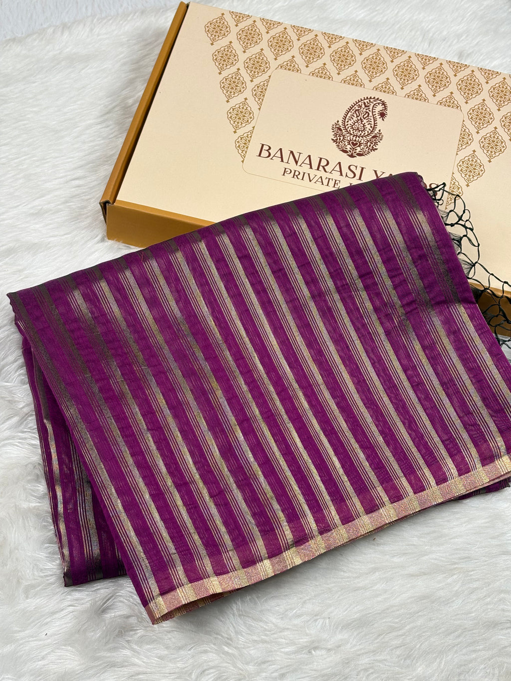 A folded purple Banarasi saree with vertical gold stripes, displayed next to its branded gift box on a white faux fur surface.