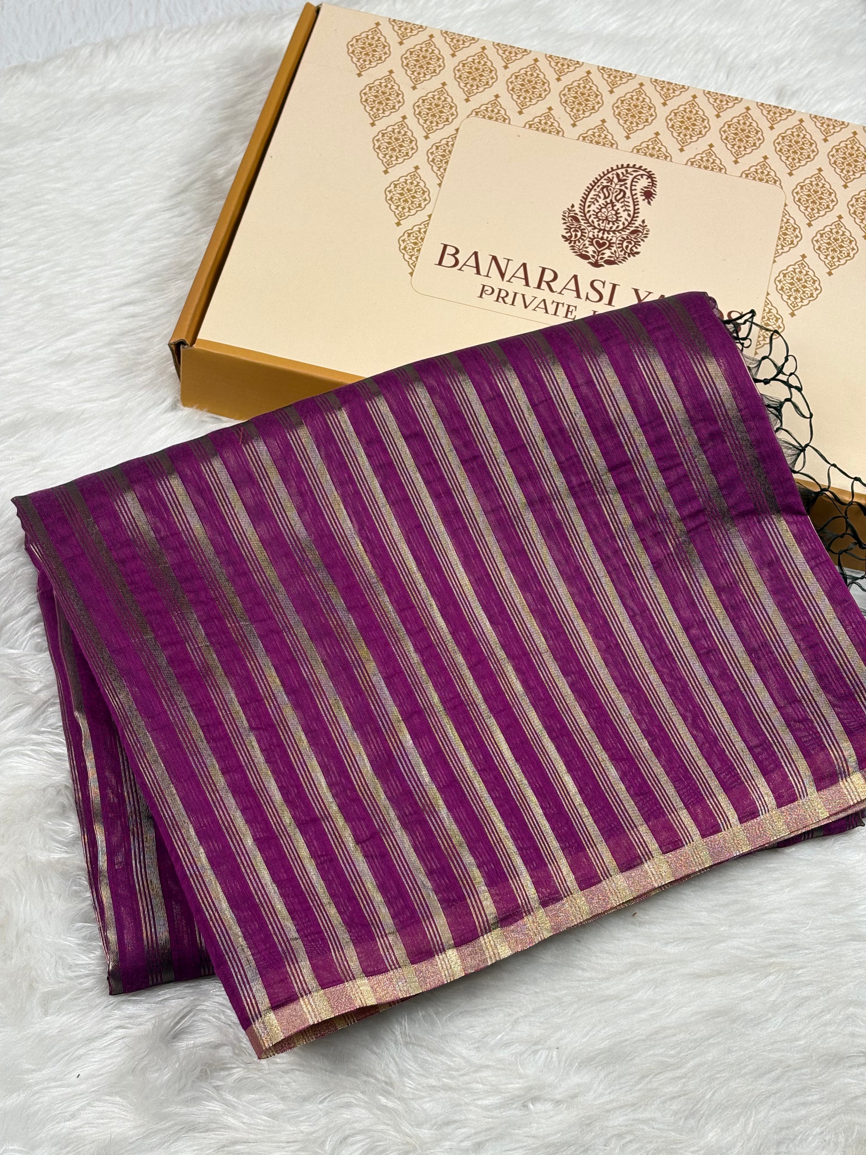 A folded purple Banarasi saree with vertical gold stripes, displayed next to its branded gift box on a white faux fur surface.