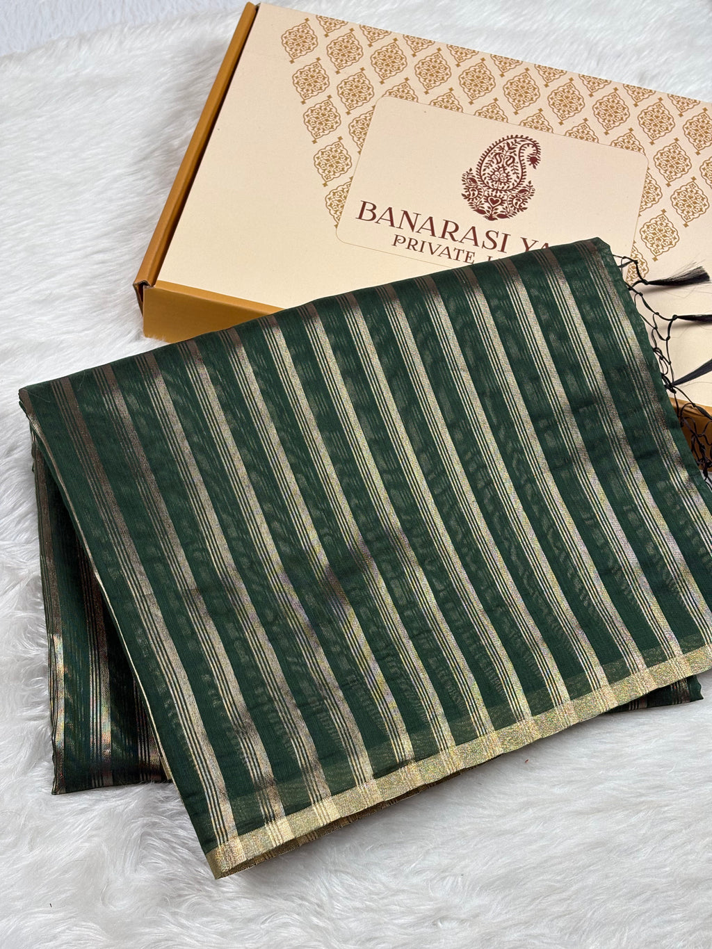 A folded dark green Banarasi saree with vertical gold stripes and a gold border, displayed next to its branded gift box on a white faux fur surface.