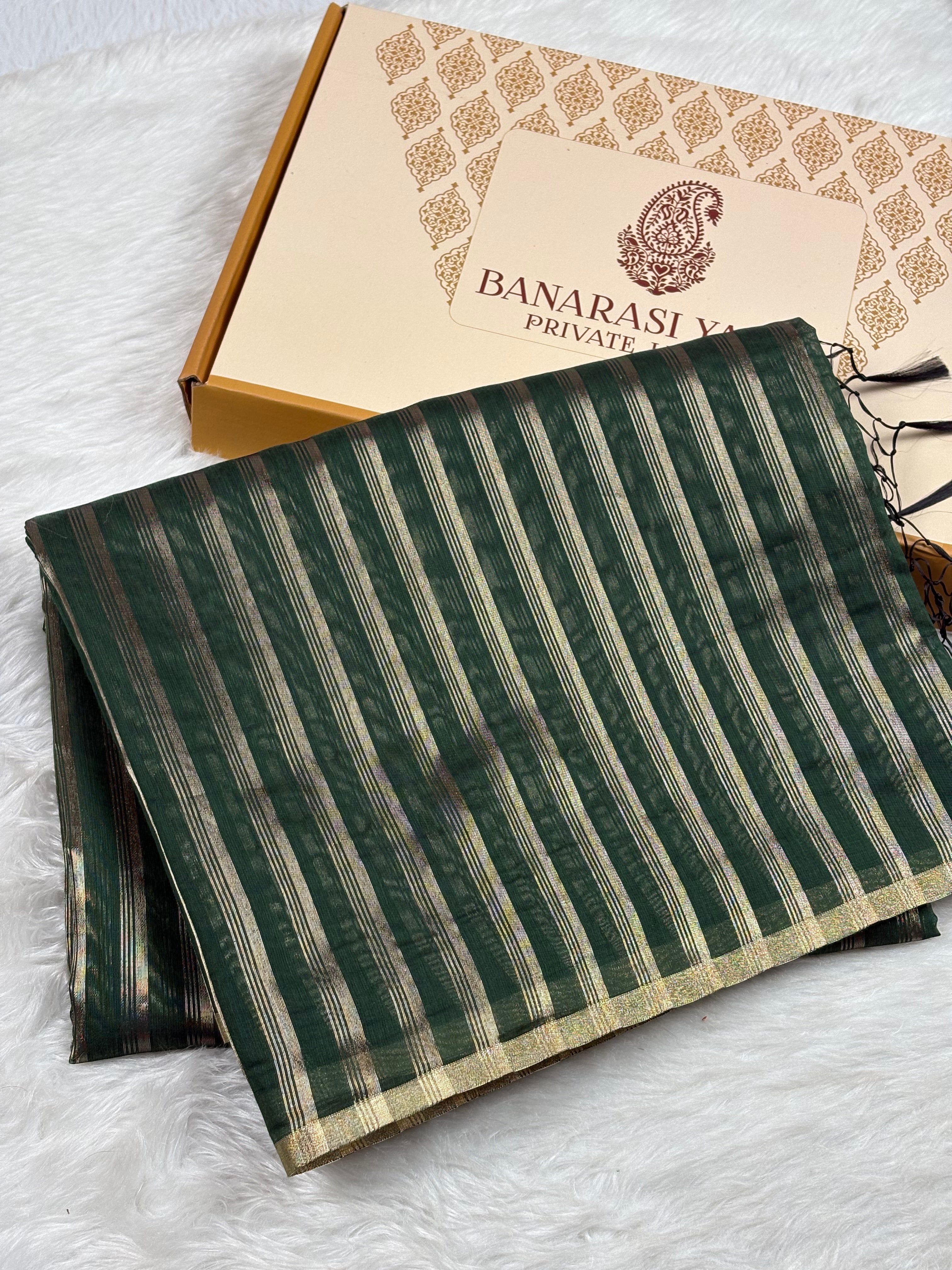 A folded dark green Banarasi saree with vertical gold stripes and a gold border, displayed next to its branded gift box on a white faux fur surface.