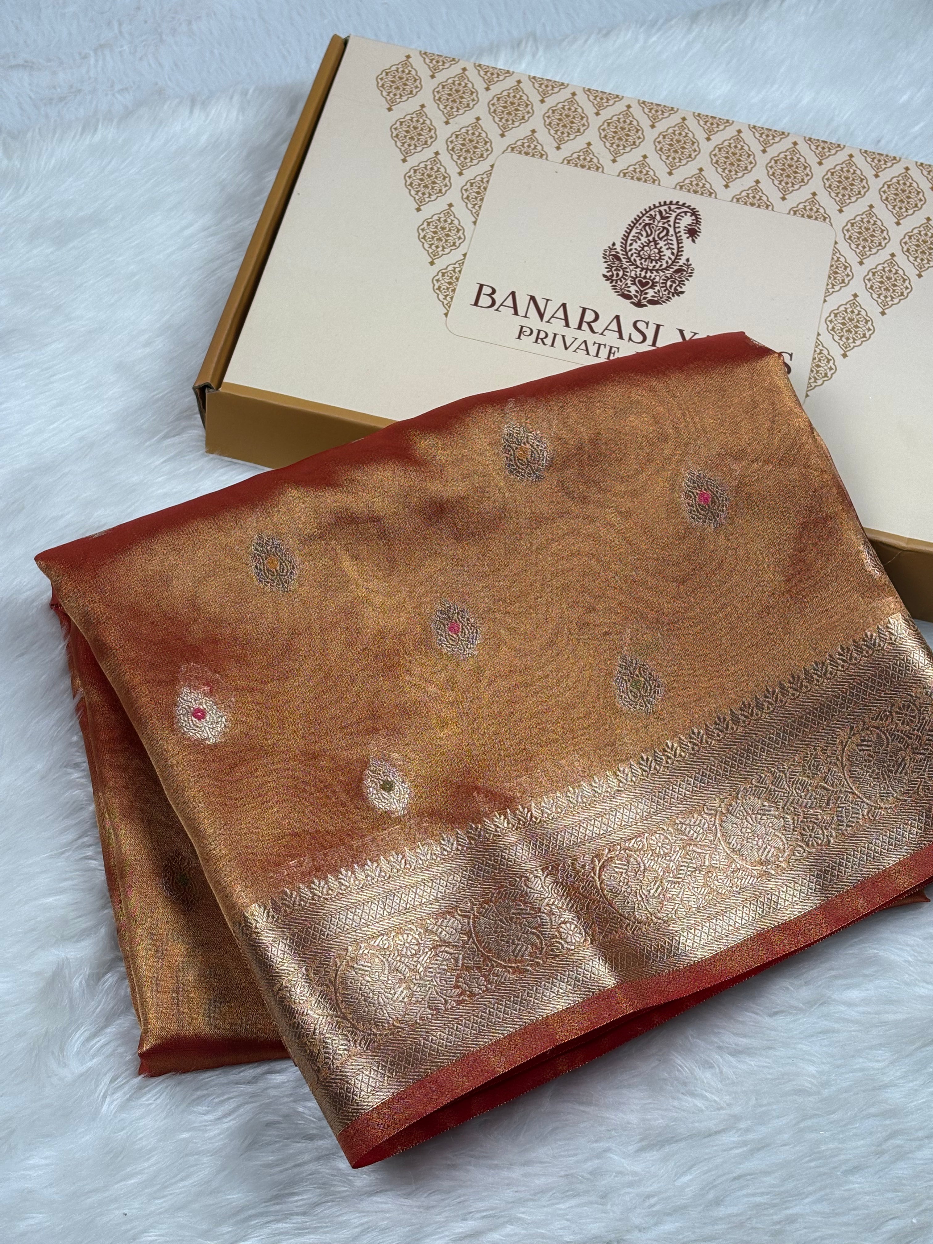 A folded Banarasi silk saree in a warm golden-orange hue with gold zari motifs and a red border, displayed next to its branded presentation box on a white faux fur surface.