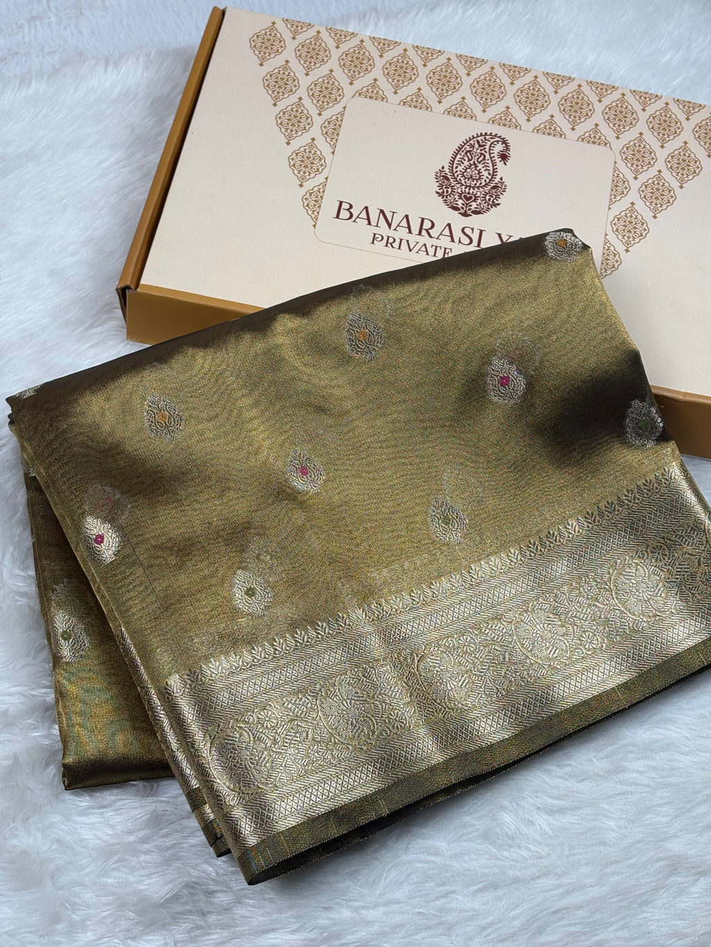 A folded Banarasi silk saree in olive green with intricate gold zari weaving and small mina accents, displayed next to its branded packaging box on a white faux fur surface.