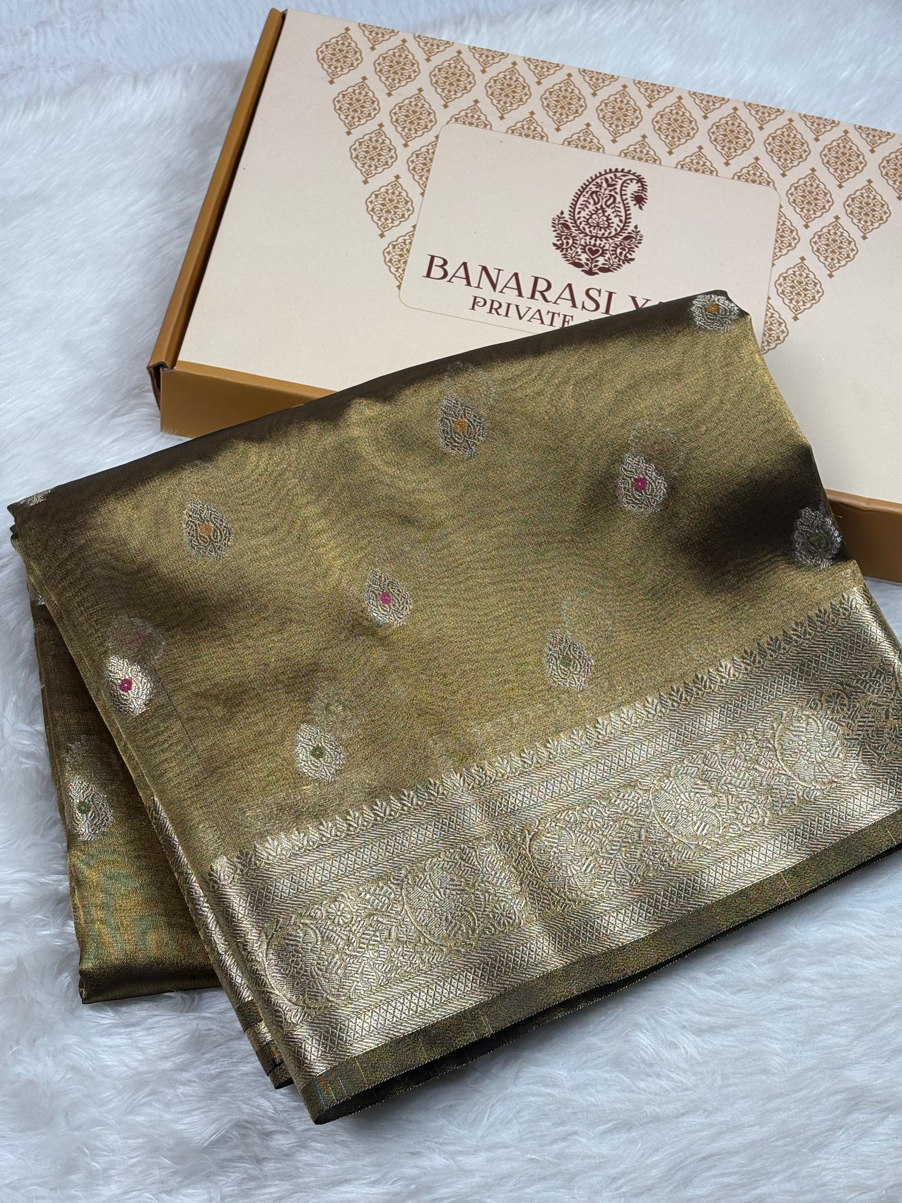 A folded Banarasi silk saree in olive green with intricate gold zari weaving and small mina accents, displayed next to its branded packaging box on a white faux fur surface.