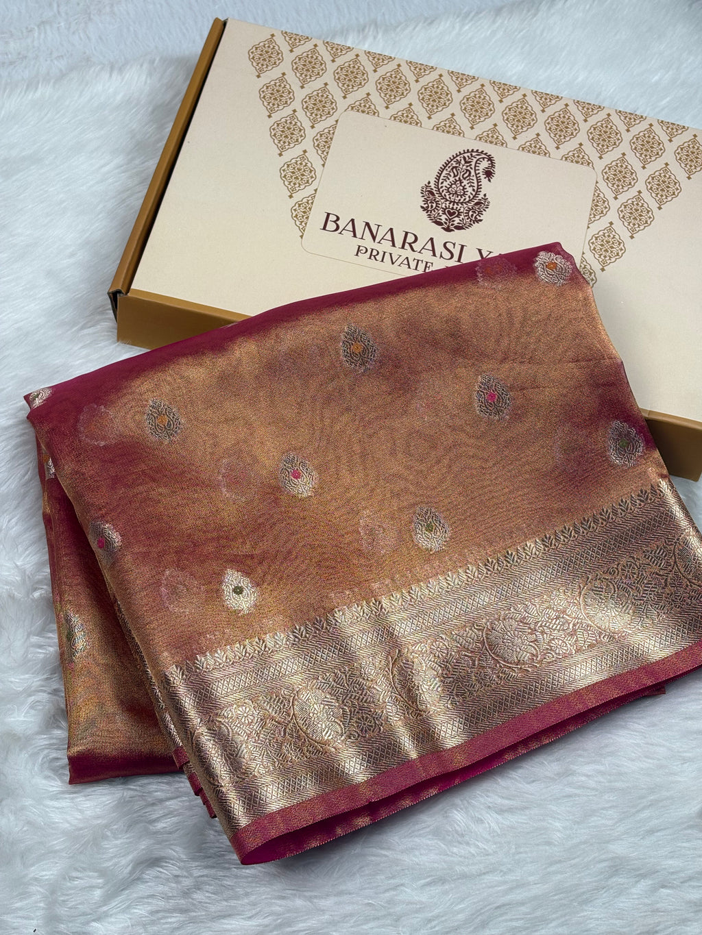 A folded Banaras silk saree in a warm coppery-brown tone with gold zari weaving and small mina accents, displayed next to its packaging box on a white faux fur surface.