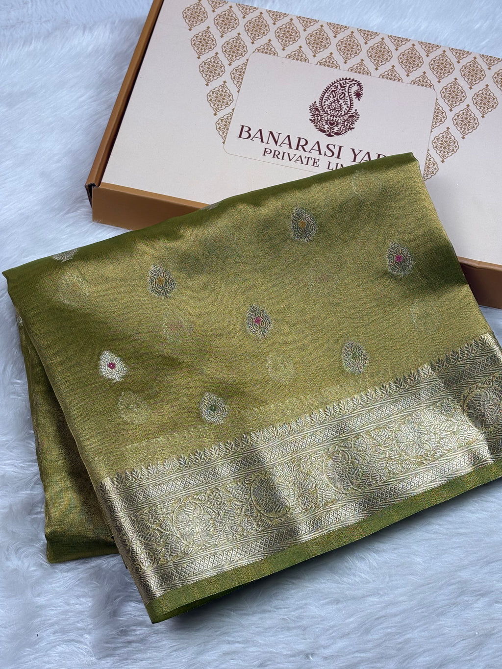 An olive green silk saree with a gold zari border and motifs, folded next to its branded Banarasi Yards Private Limited gift box on a white faux fur surface.