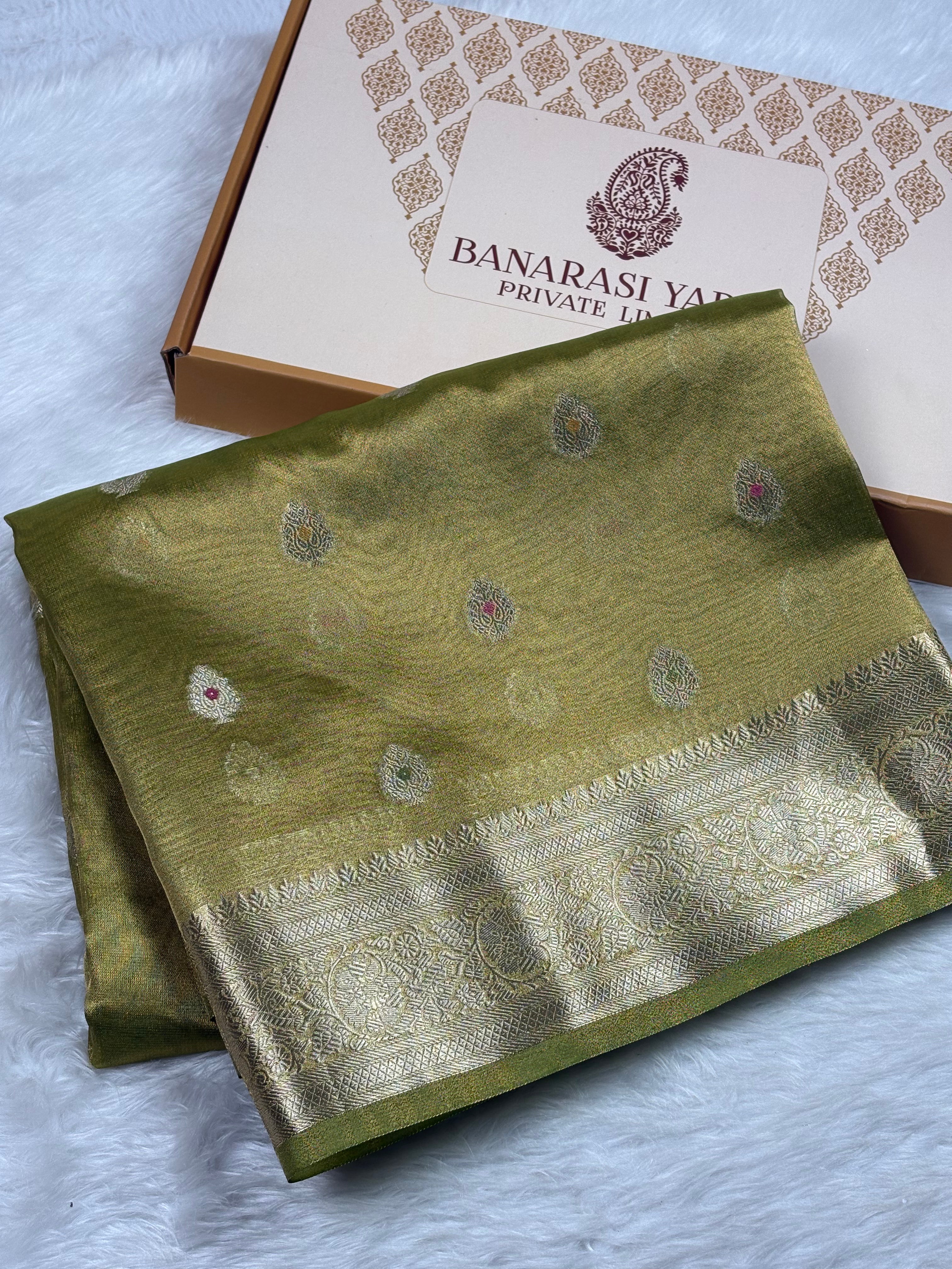 An olive green silk saree with a gold zari border and motifs, folded next to its branded Banarasi Yards Private Limited gift box on a white faux fur surface.