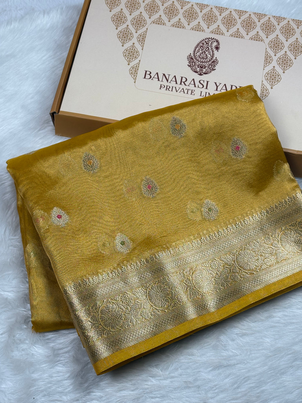 A folded mustard yellow Banarasi silk saree with a wide gold zari border and small floral mina motifs, displayed next to its branded packaging box on a white faux fur surface.