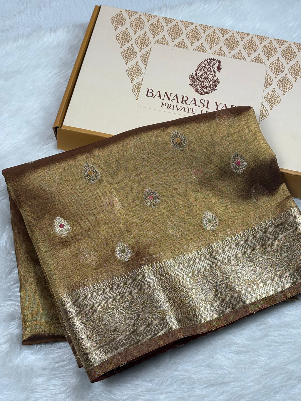 A folded Banarasi silk saree in a gold and bronze color with intricate zari weaving, displayed next to its branded packaging box on a white faux fur surface.