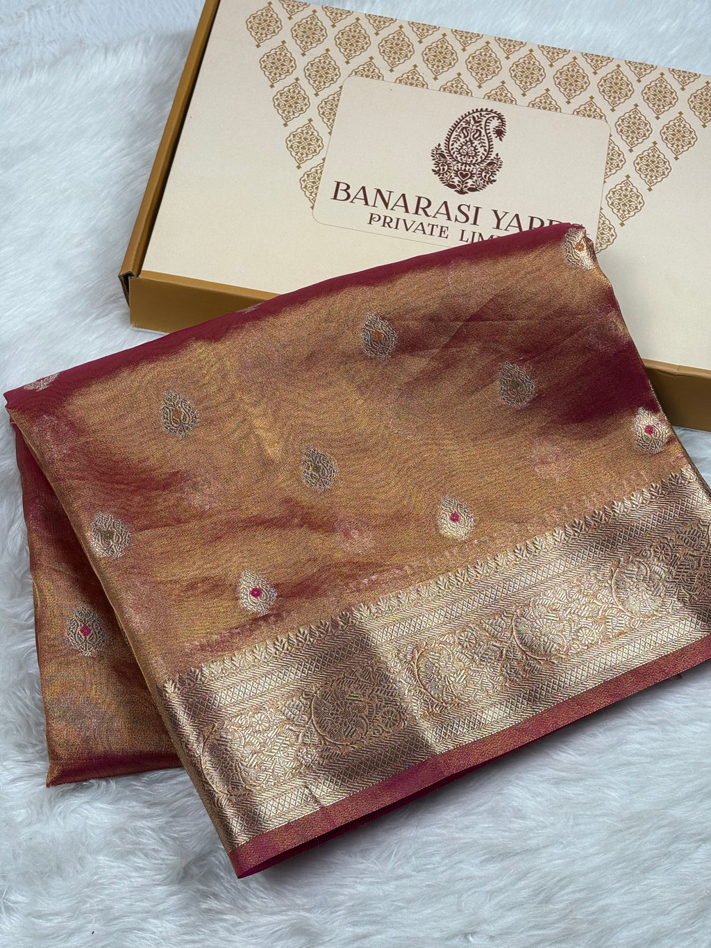 A folded gold and red Banarasi silk saree with intricate zari weaving, displayed next to its branded packaging box on a white faux fur surface.