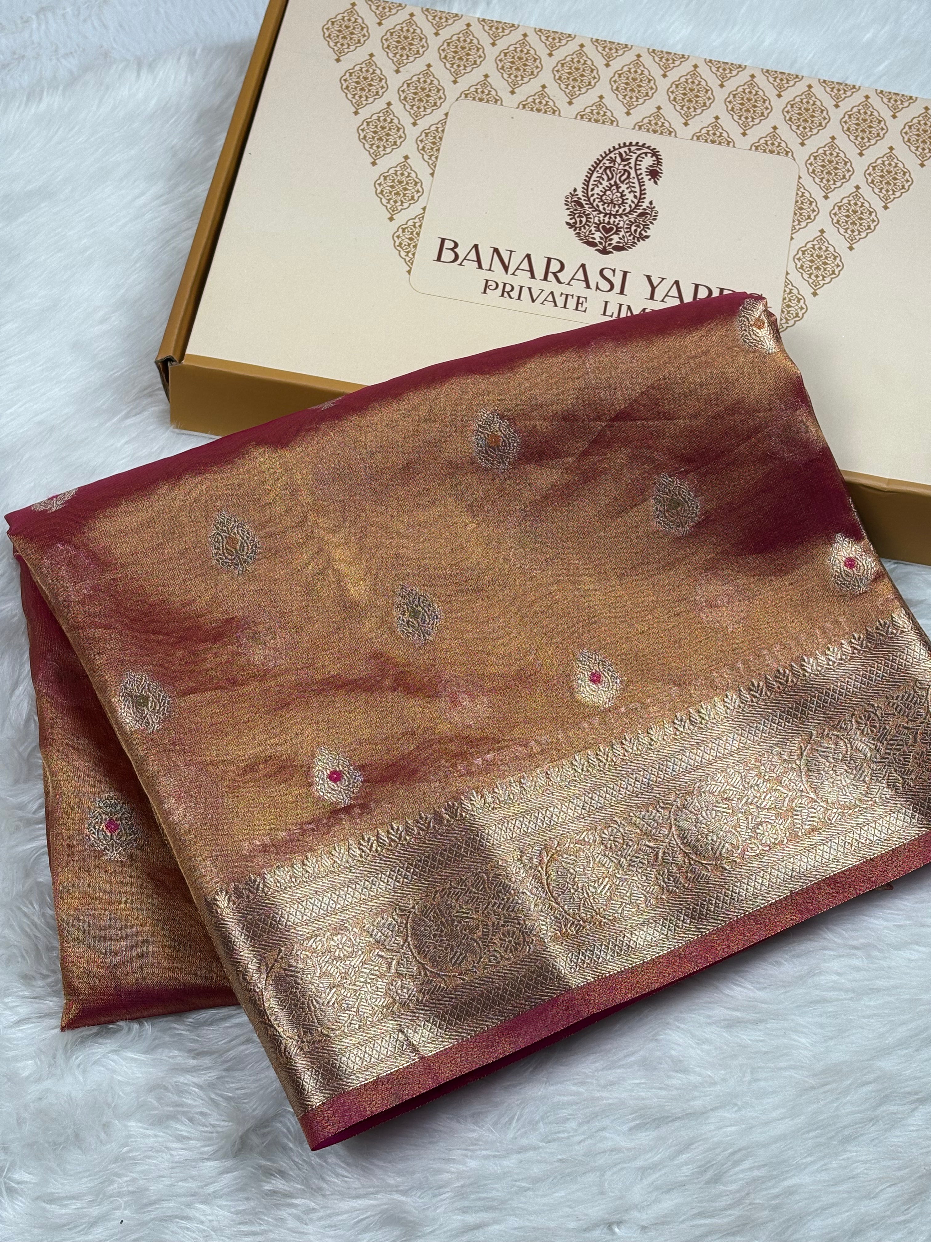 A folded gold and red Banarasi silk saree with intricate zari weaving, displayed next to its branded packaging box on a white faux fur surface.
