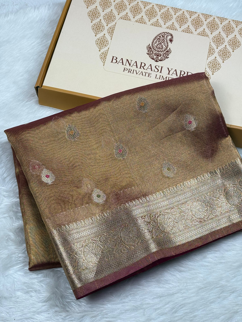 A folded Banarasi silk saree in a warm brown and gold tone with traditional zari weaving and a deep maroon border, displayed next to its branded packaging box on a white faux fur surface.
