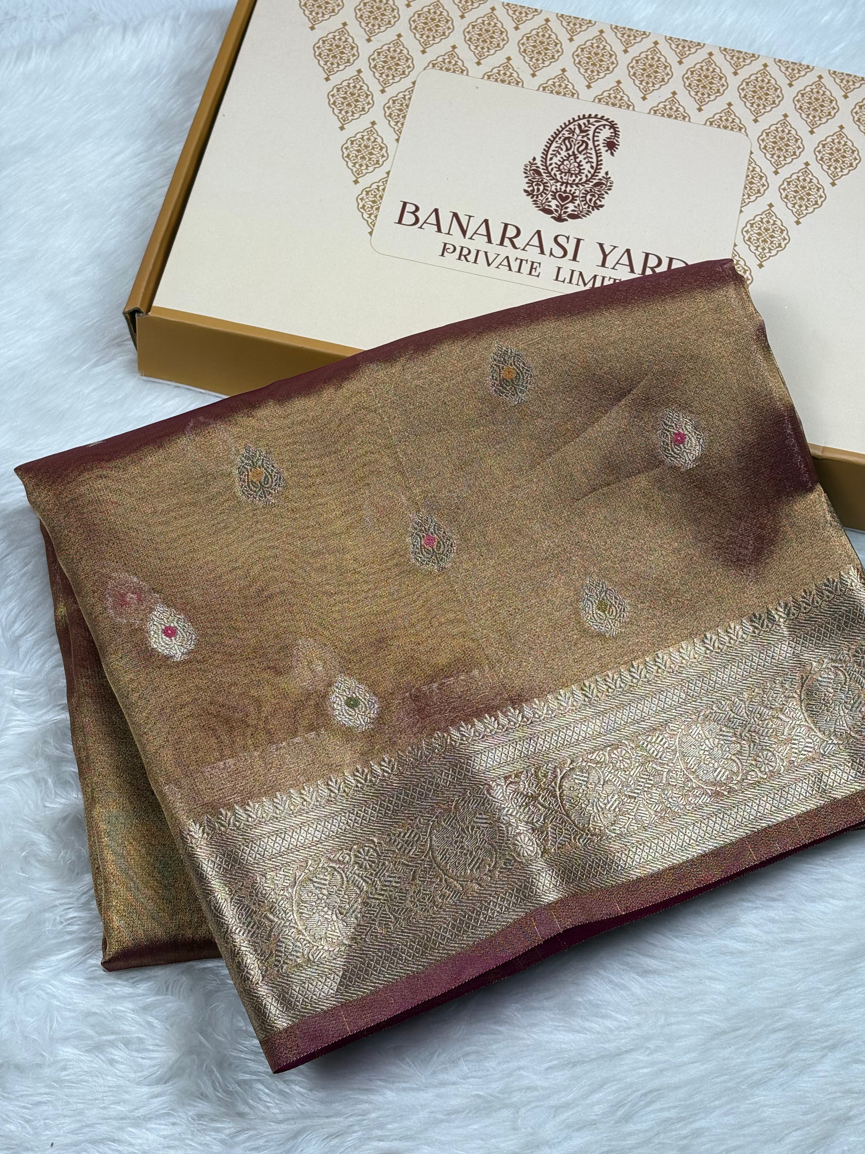 A folded Banarasi silk saree in a warm brown and gold tone with traditional zari weaving and a deep maroon border, displayed next to its branded packaging box on a white faux fur surface.