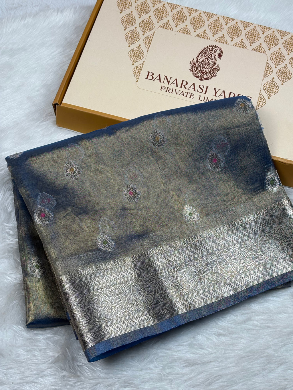 A folded Banarasi silk saree with a blue and gold body and a zari border, displayed next to its branded packaging box on a white faux fur surface.