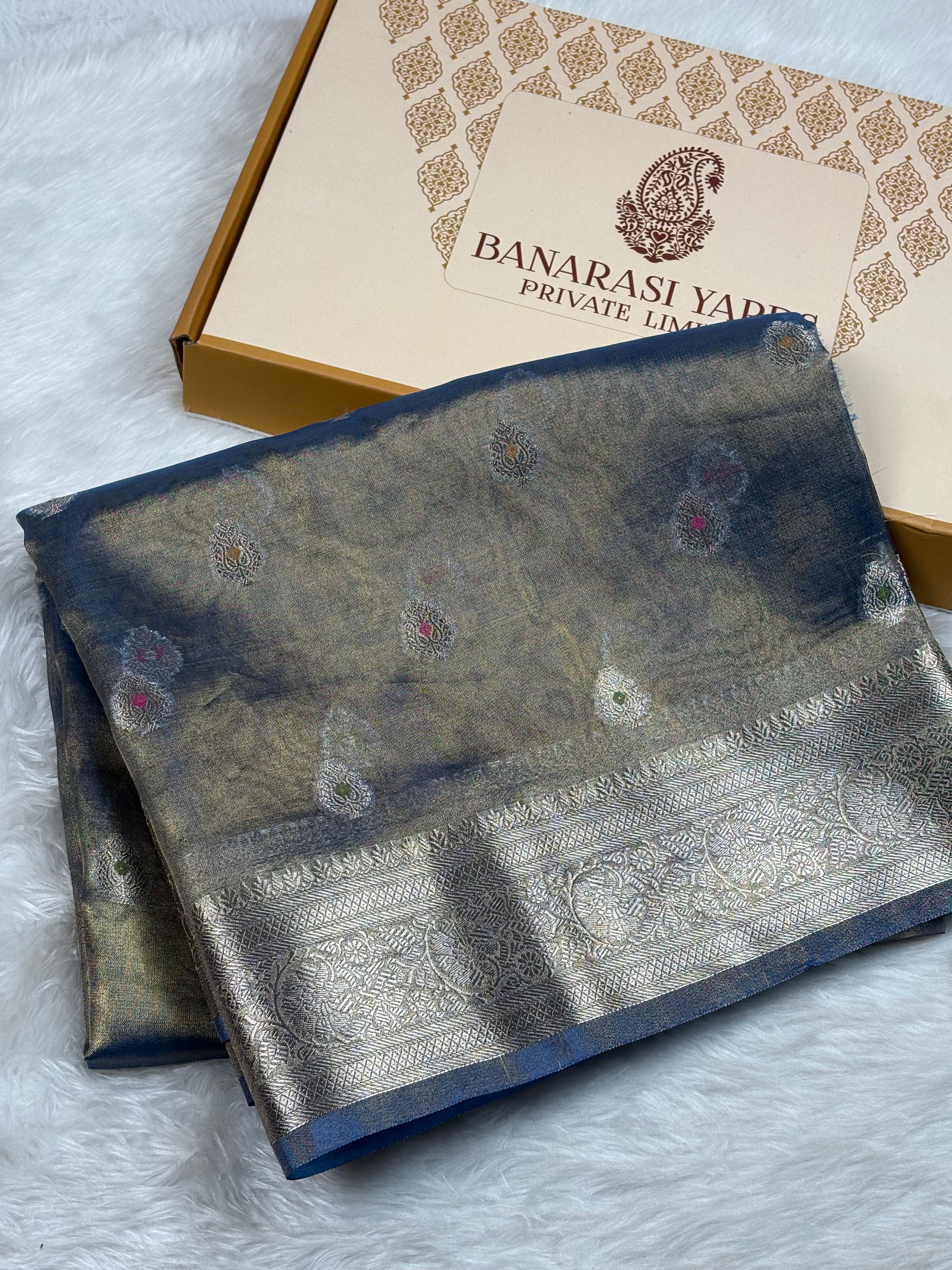 A folded Banarasi silk saree with a blue and gold body and a zari border, displayed next to its branded packaging box on a white faux fur surface.