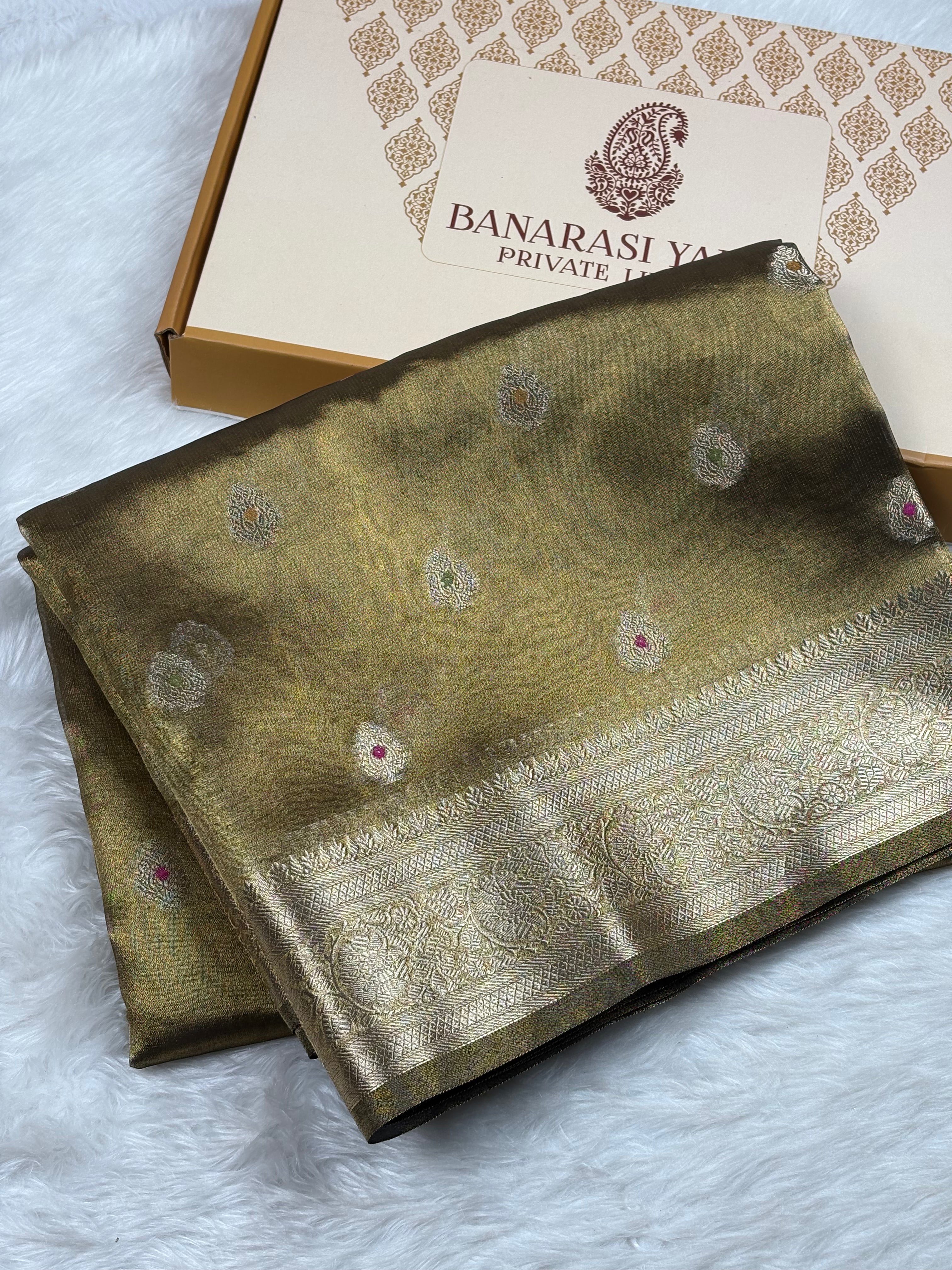 An olive green Banarasi silk saree with gold zari weaving and small floral mina motifs, folded next to its branded packaging box on a white faux fur surface.