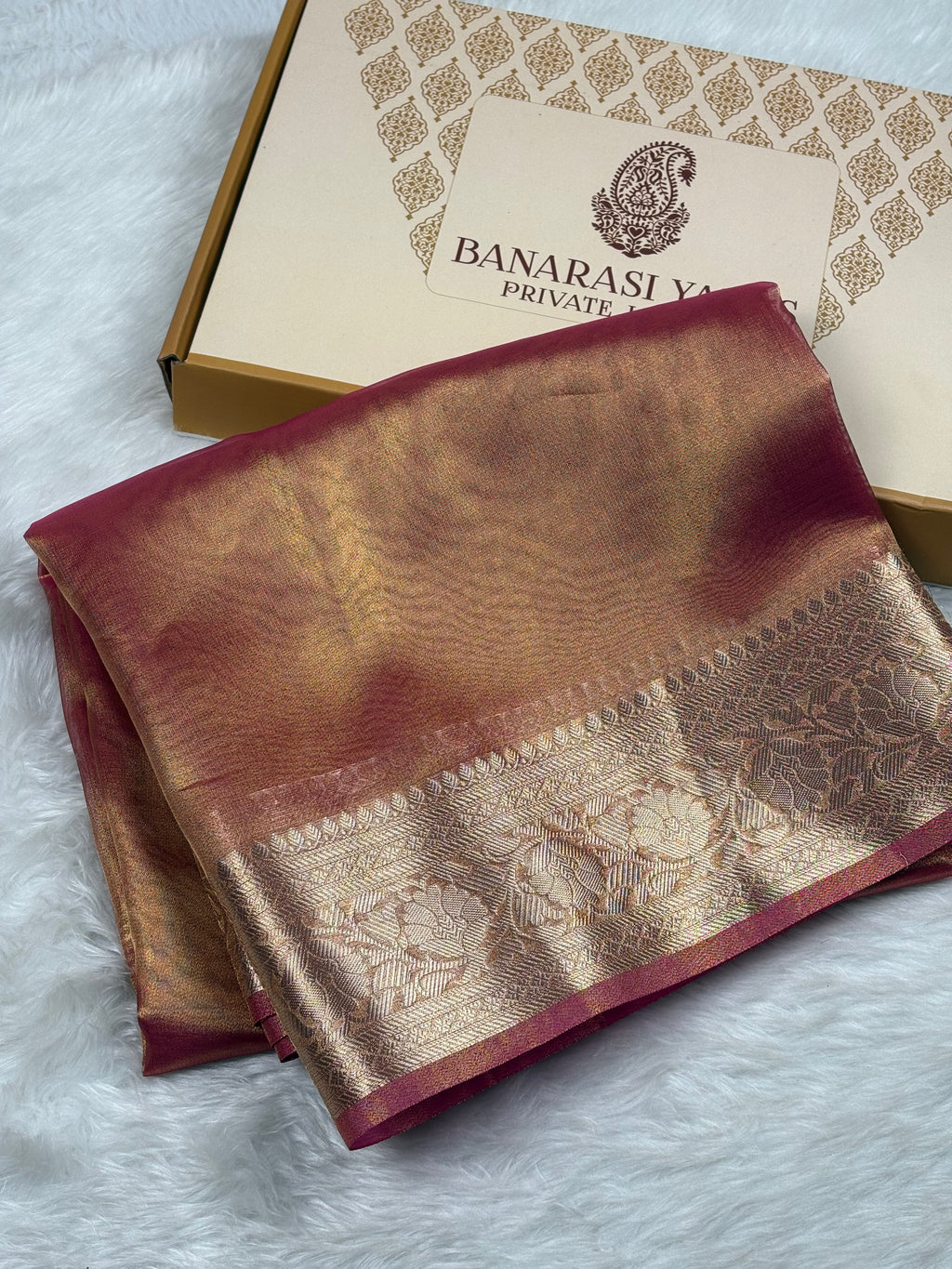 A folded Banarasi silk saree with a deep maroon border and a shimmering gold zari pattern, displayed next to its branded gift box on a white faux fur surface.