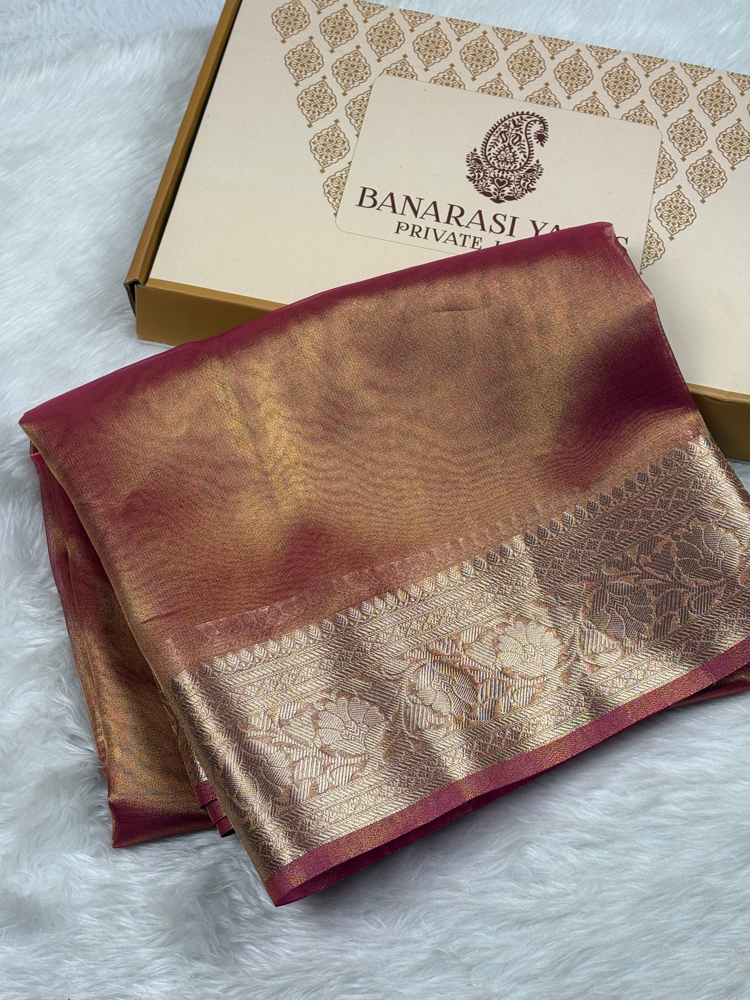 A folded Banarasi silk saree with a deep maroon border and a shimmering gold zari pattern, displayed next to its branded gift box on a white faux fur surface.