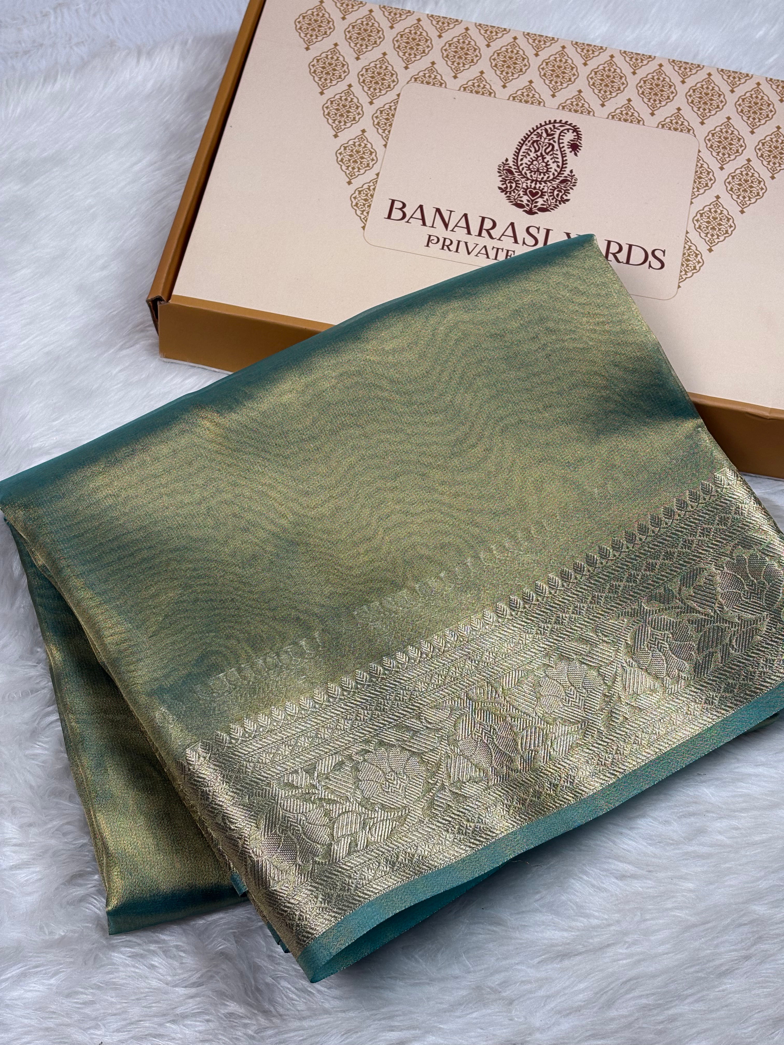 A folded green and gold Banarasi silk saree with a woven border, displayed next to its branded gift box on a white faux fur surface.