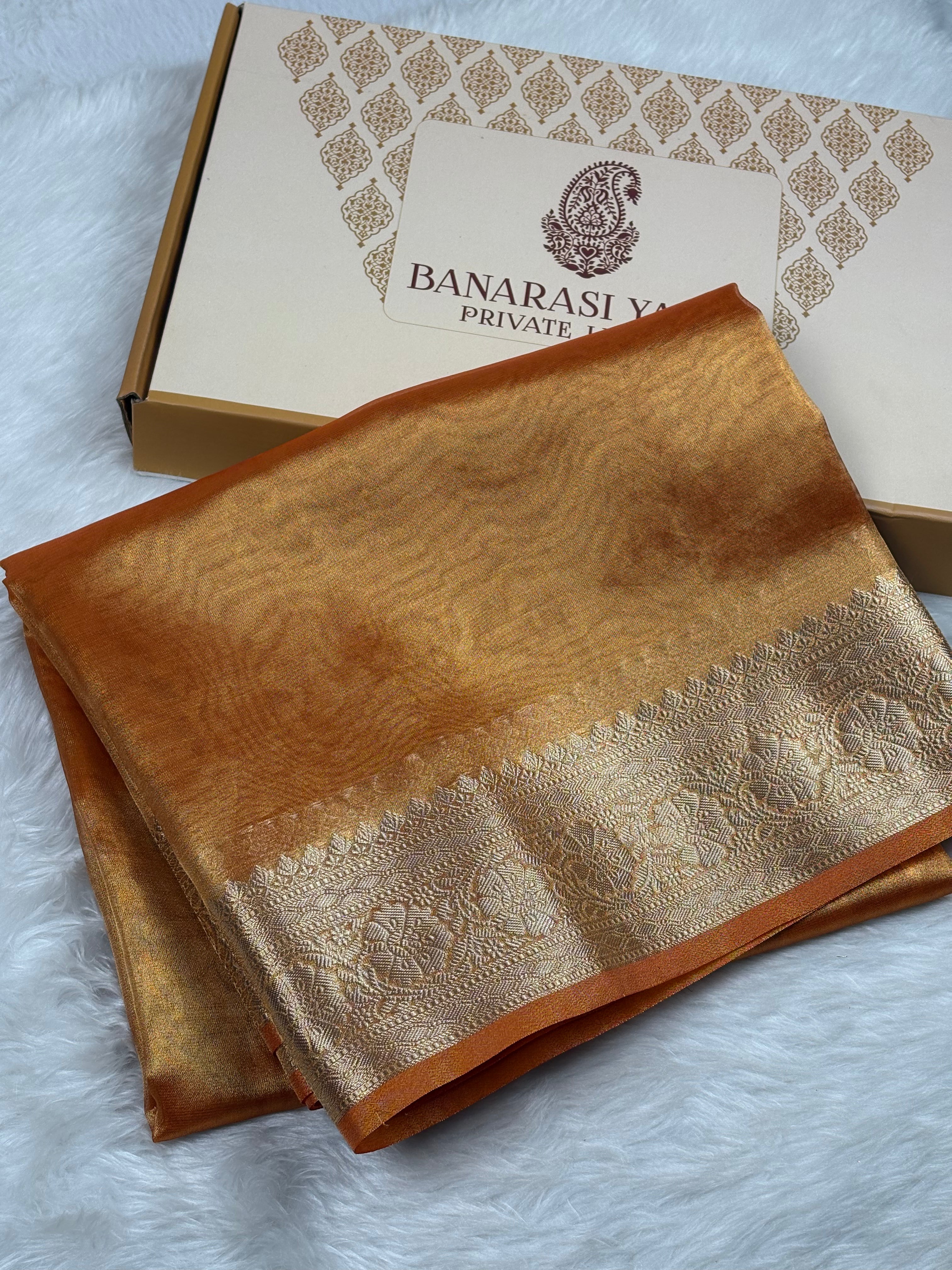 A folded golden-brown Banarasi silk saree with a zari border, displayed next to its branded packaging box on a white faux fur surface.