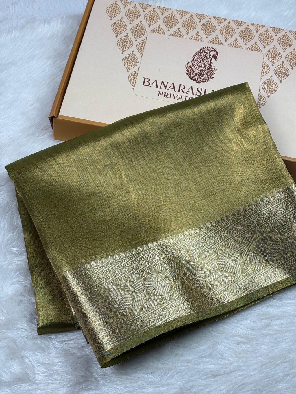 An olive green silk saree with a wide gold zari border, folded and displayed next to its branded 'Banarasi Yards Private Limited' gift box on a white faux fur surface.