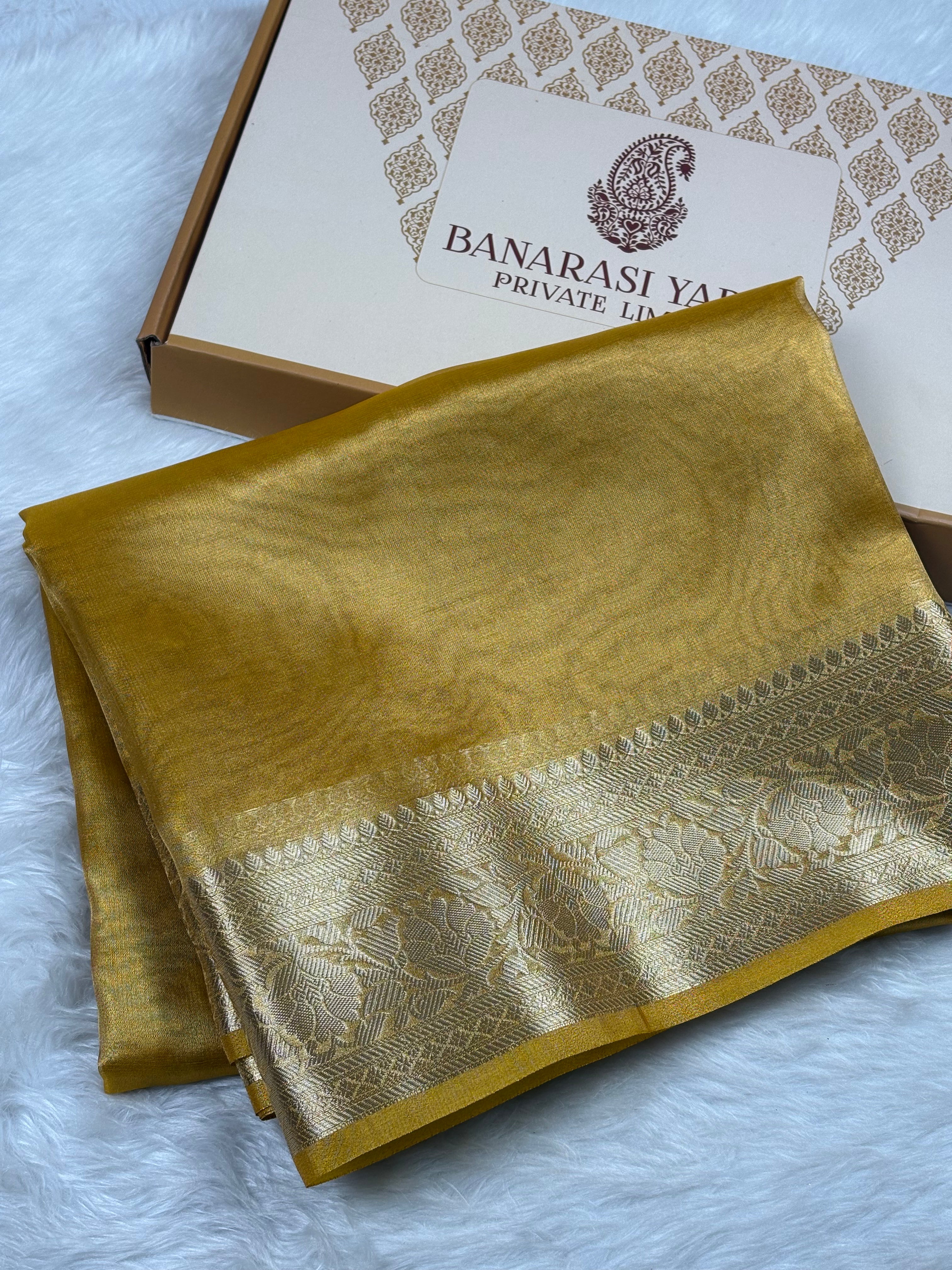 A folded mustard yellow silk saree with a wide gold zari border, displayed next to its branded packaging box on a white faux fur surface.