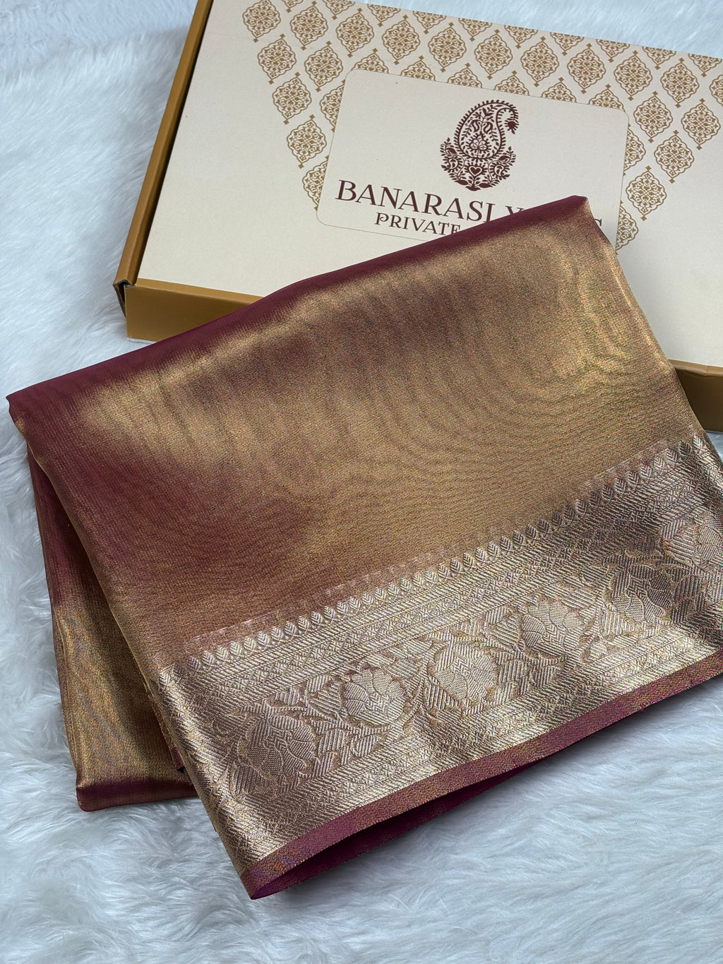 A folded Banarasi silk saree in a gold and deep maroon color, displayed next to its branded packaging box on a white faux fur surface.
