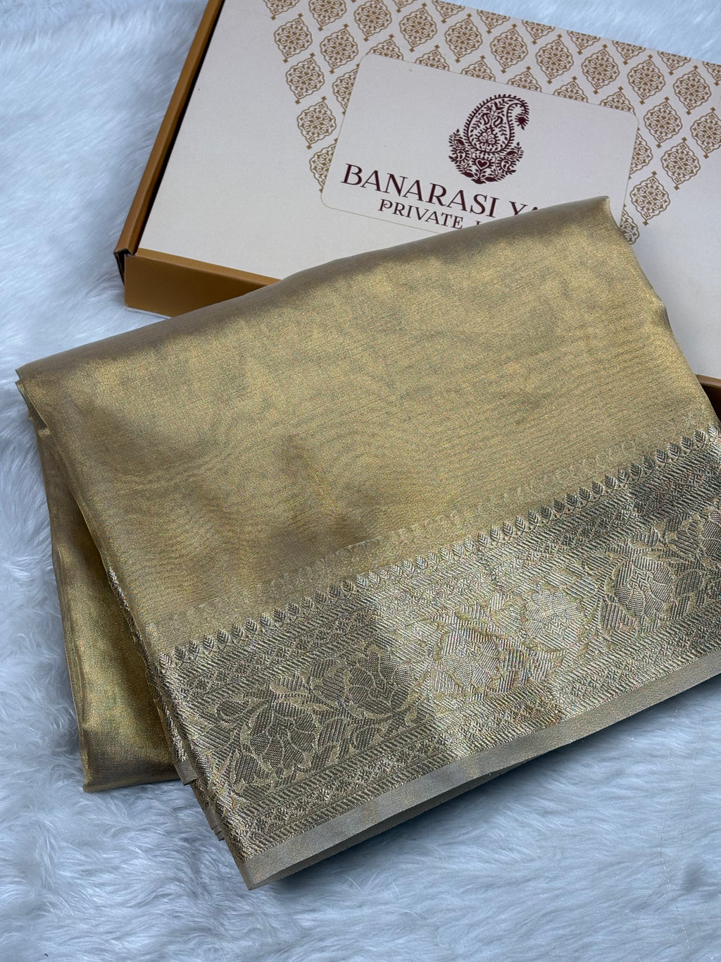 A folded gold-colored Banarasi silk saree with a detailed floral zari border, displayed next to its branded packaging box on a white faux fur surface.