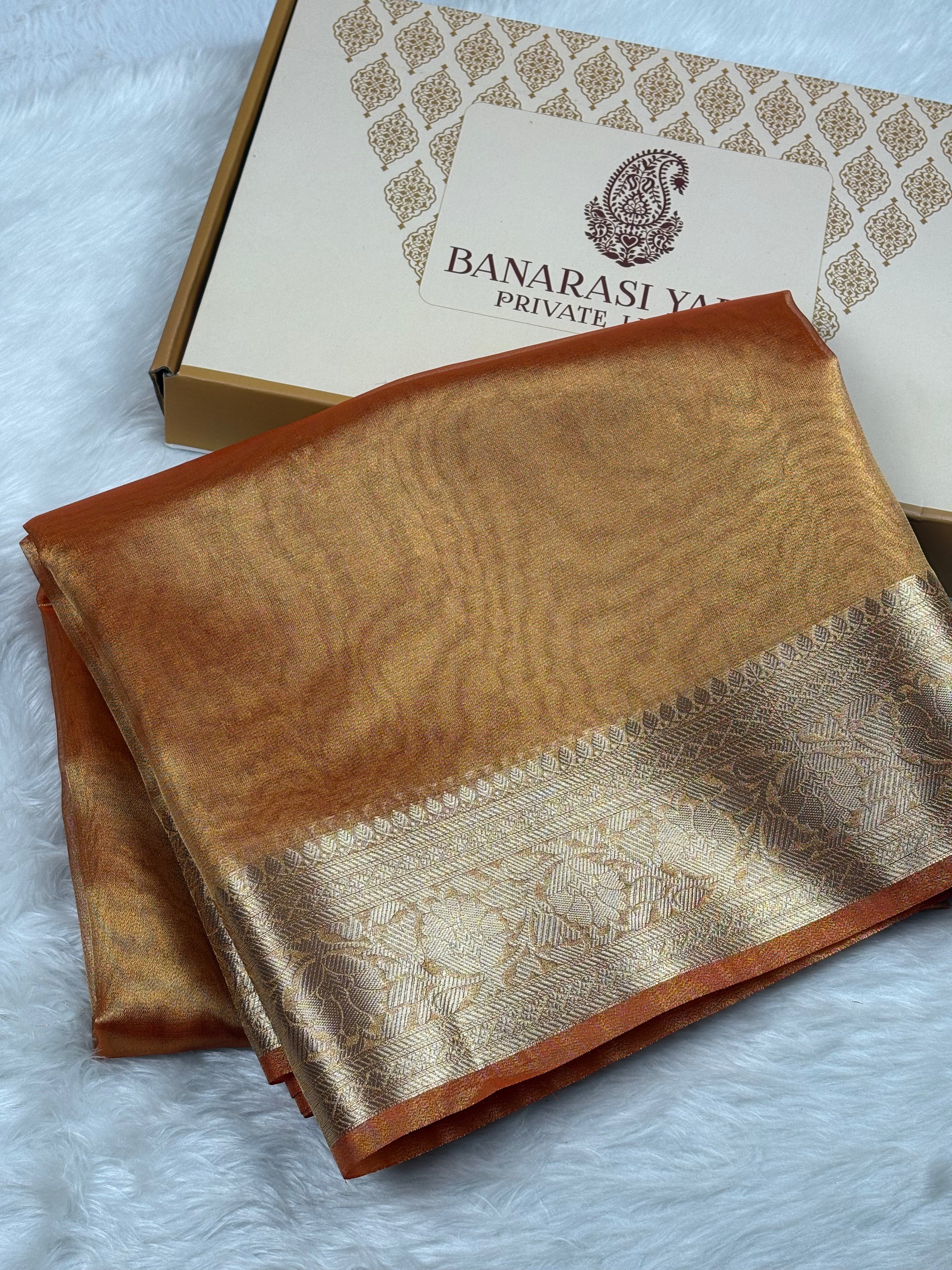 A folded golden-brown Banarasi silk saree with a zari border, displayed next to its branded packaging box on a white faux fur surface.