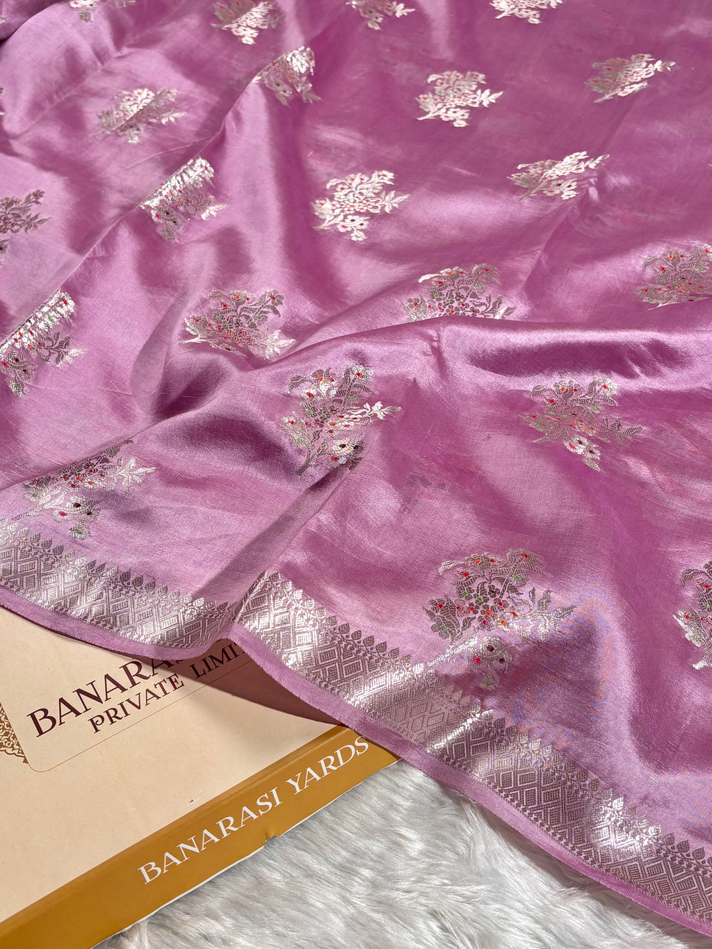 A close-up of a pink Banarasi pure katan silk saree with silver floral tilfi mina motifs and a silver border, draped over a paper label that reads 'BANARASI YARDS'.