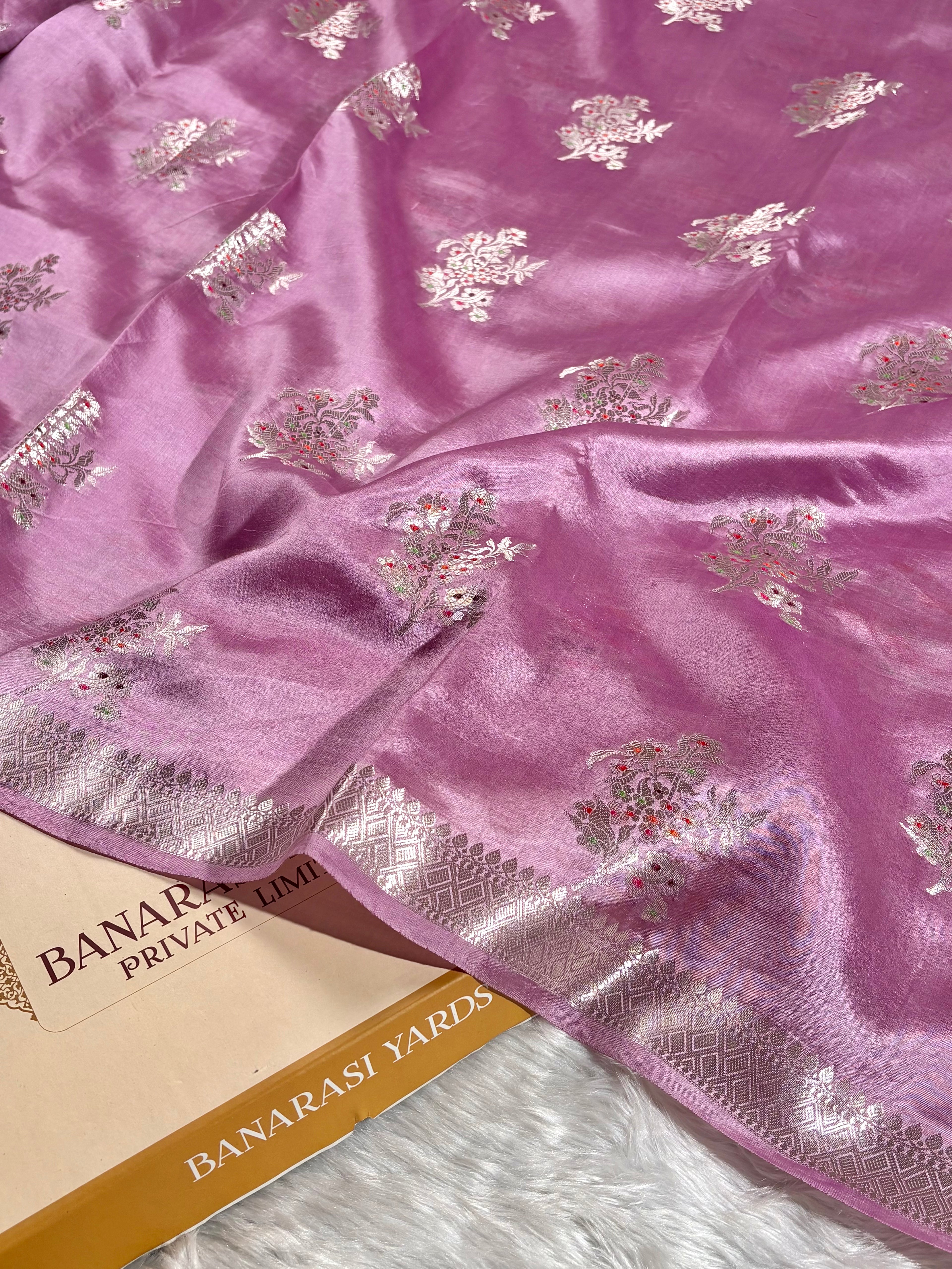A close-up of a pink Banarasi pure katan silk saree with silver floral tilfi mina motifs and a silver border, draped over a paper label that reads 'BANARASI YARDS'.