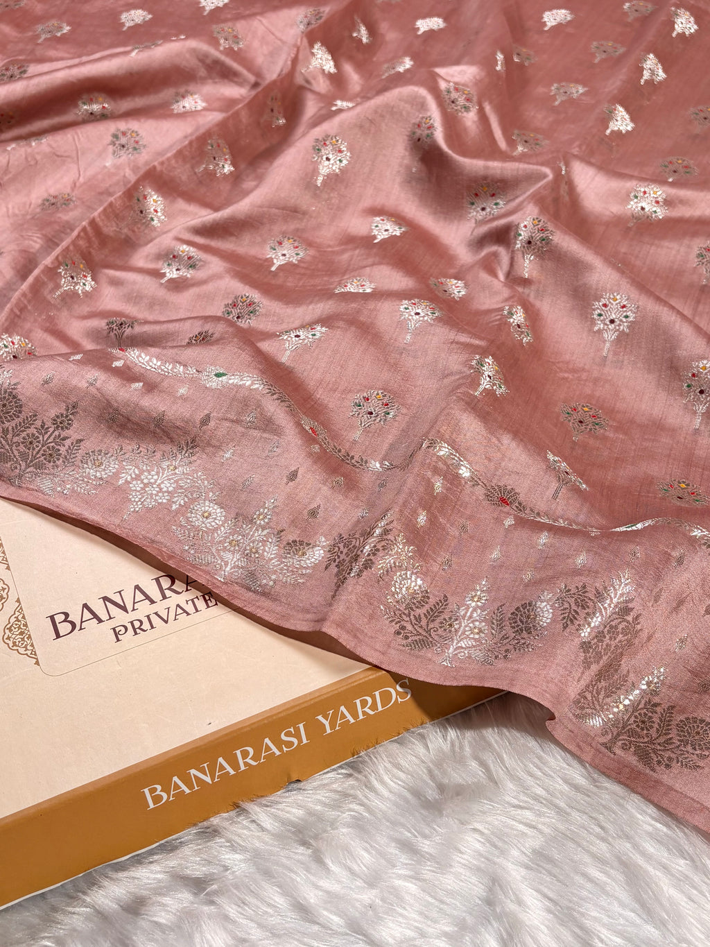 A dusty pink Banarasi silk saree with silver floral and tilfi mina weaving, draped elegantly over a Banarasi Yards Private Limited box on a white faux fur surface.