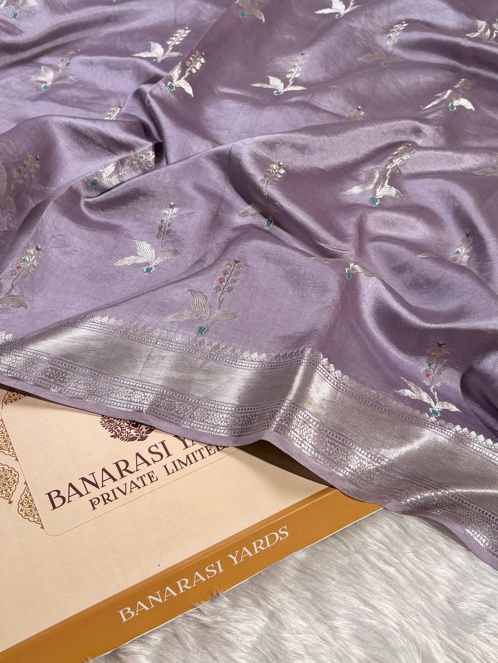 A lavender purple Banarasi silk saree with silver zari weaving of floral tilfi mina motifs, draped elegantly over a Banarasi Yards box on a white faux fur surface.